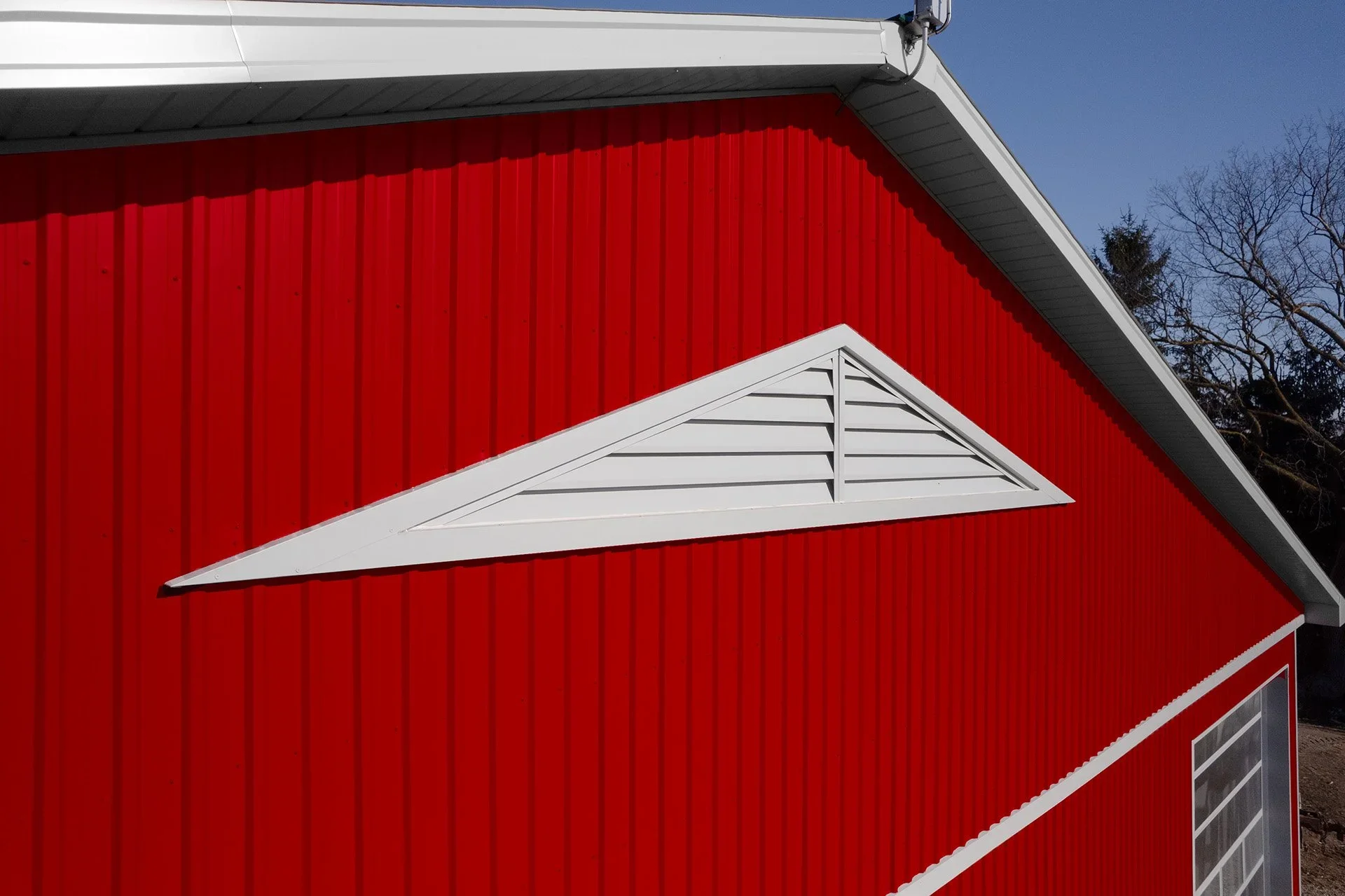Close-up of a red building with a white triangular vent and roof trim, under a clear blue sky, with bare tree branches in the background.