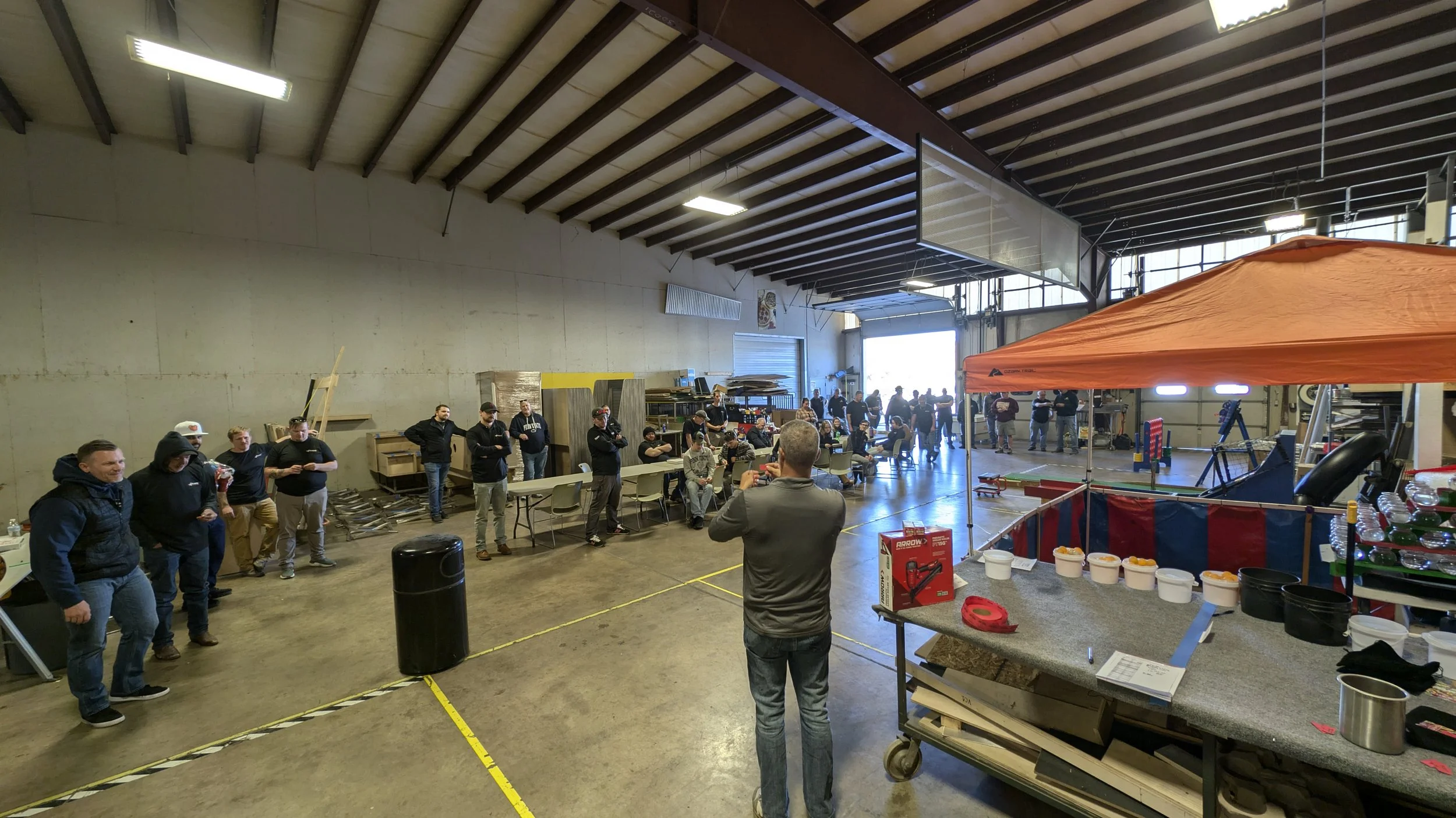 An indoor gathering space with people standing and sitting, some in a line facing left, and a man in a grey jacket giving a presentation. The area has a high ceiling with exposed beams, tables, and a booth with snacks on the right. Bright lighting an