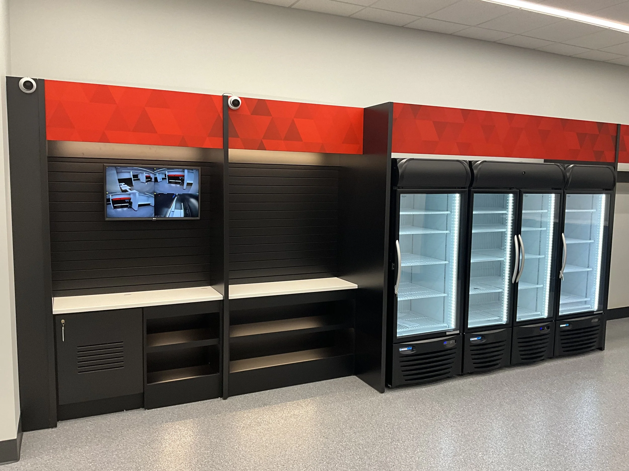 Empty refrigerated display cases and shelving unit in a store, with a monitor showing CCTV footage.