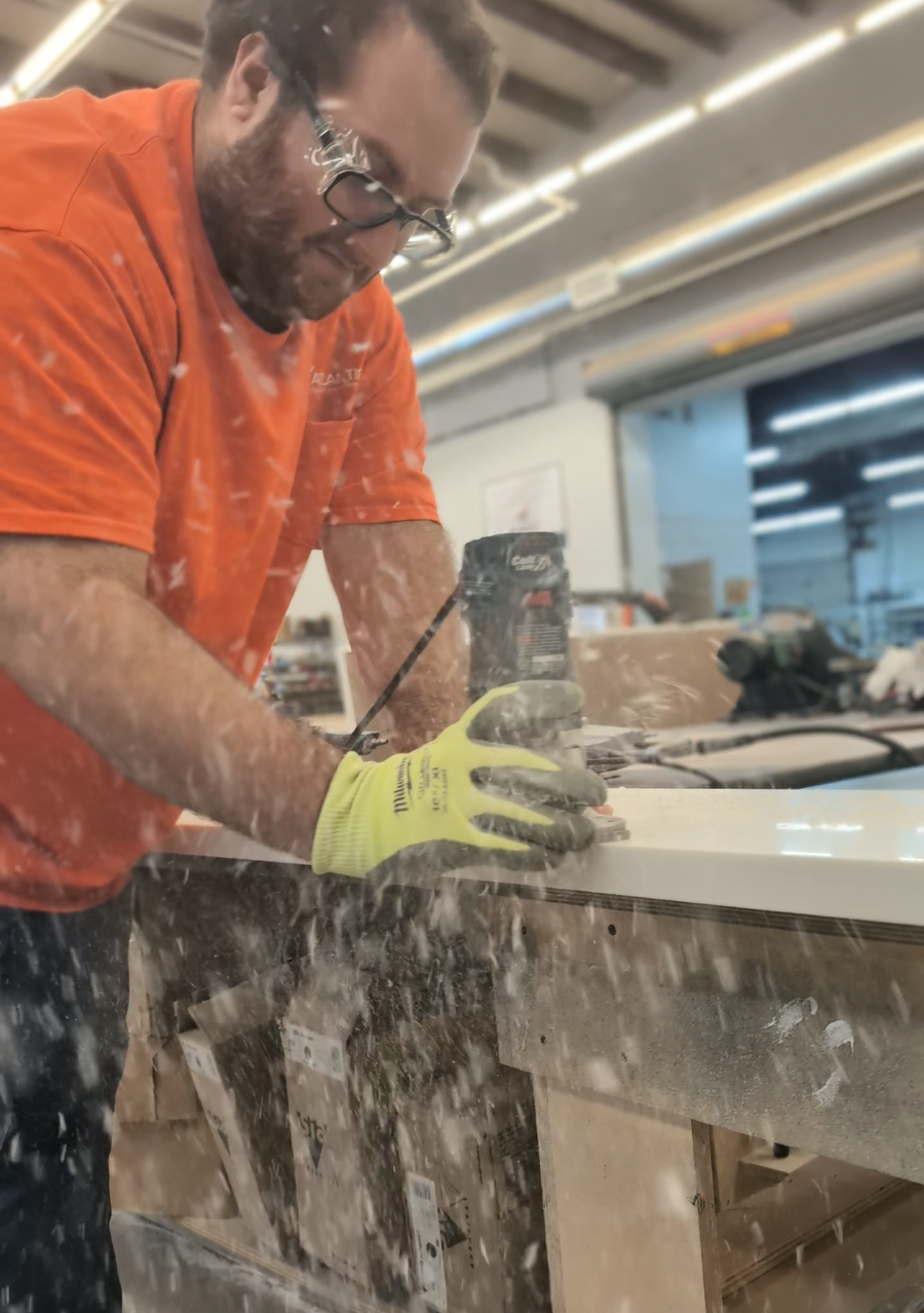 A man wearing glasses, an orange shirt, and yellow gloves using a power sander on a piece of wood in a workshop.