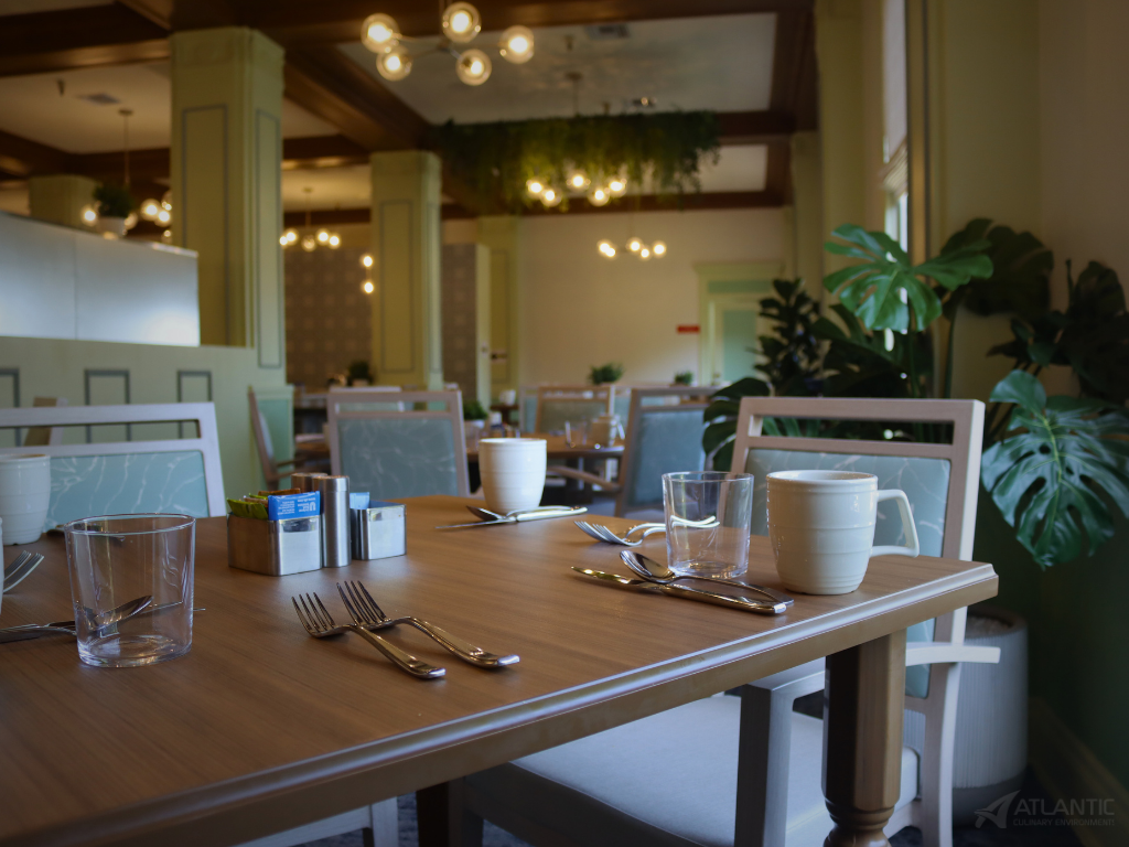 Empty restaurant table set with forks, knives, glasses, a coffee mug, a sugar packet holder, and a salt and pepper shaker, with plants and dining area in the background.