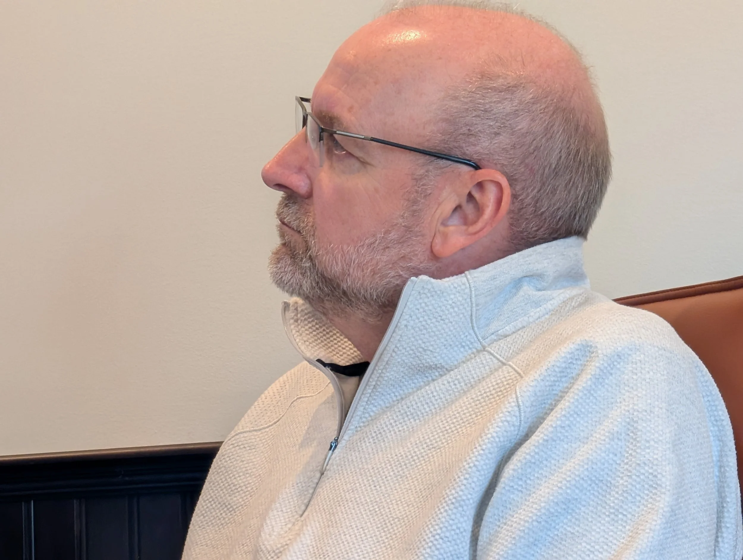 Side profile of a middle-aged man with glasses and gray hair, wearing a light-colored zip-up jacket, sitting against a plain wall.