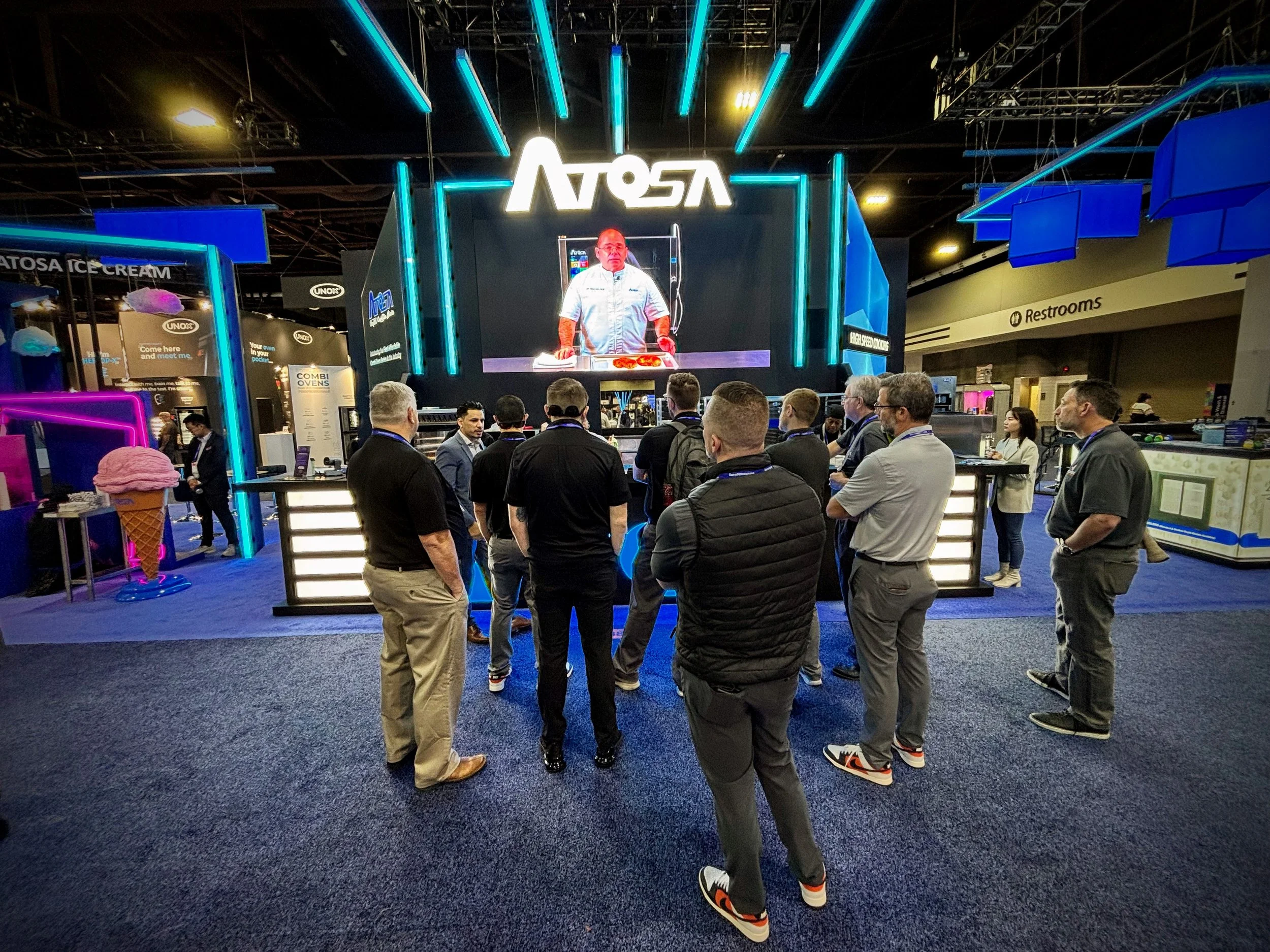 Group of people attending a presentation at a technology trade show. A large digital screen displays a chef preparing food, and the booth has vibrant blue neon lighting and signage.
