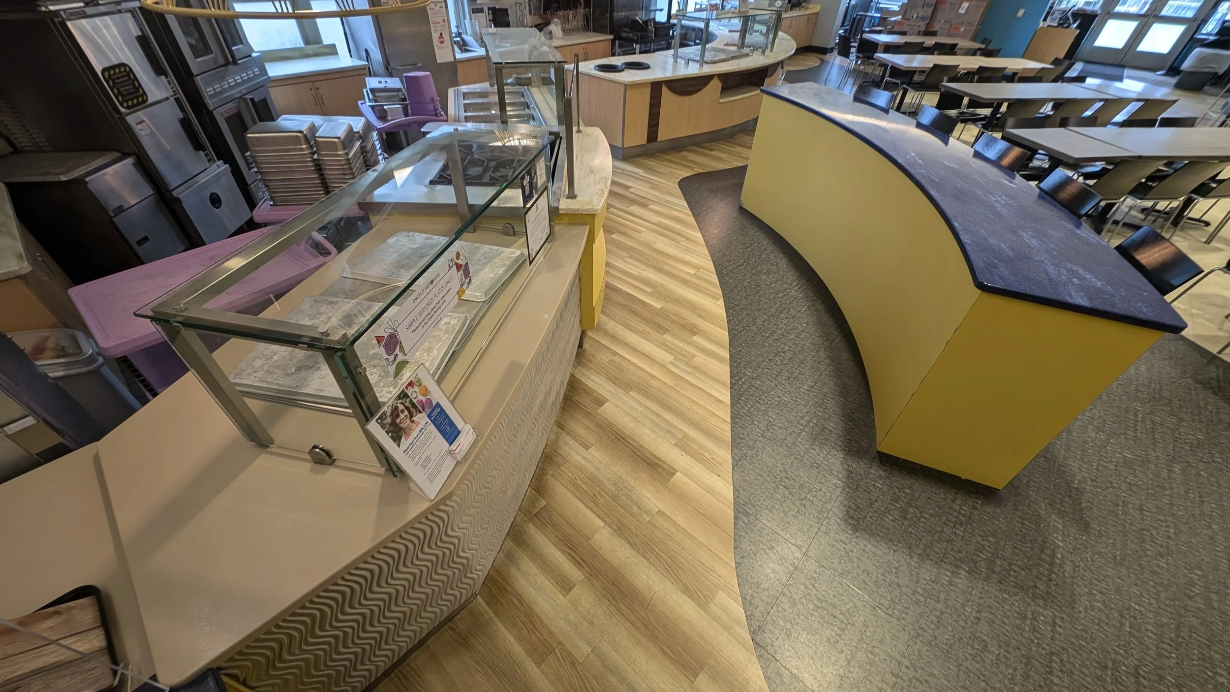 Empty cafeteria food service area with a glass display case, a curved yellow counter with a dark countertop, and tables with chairs.