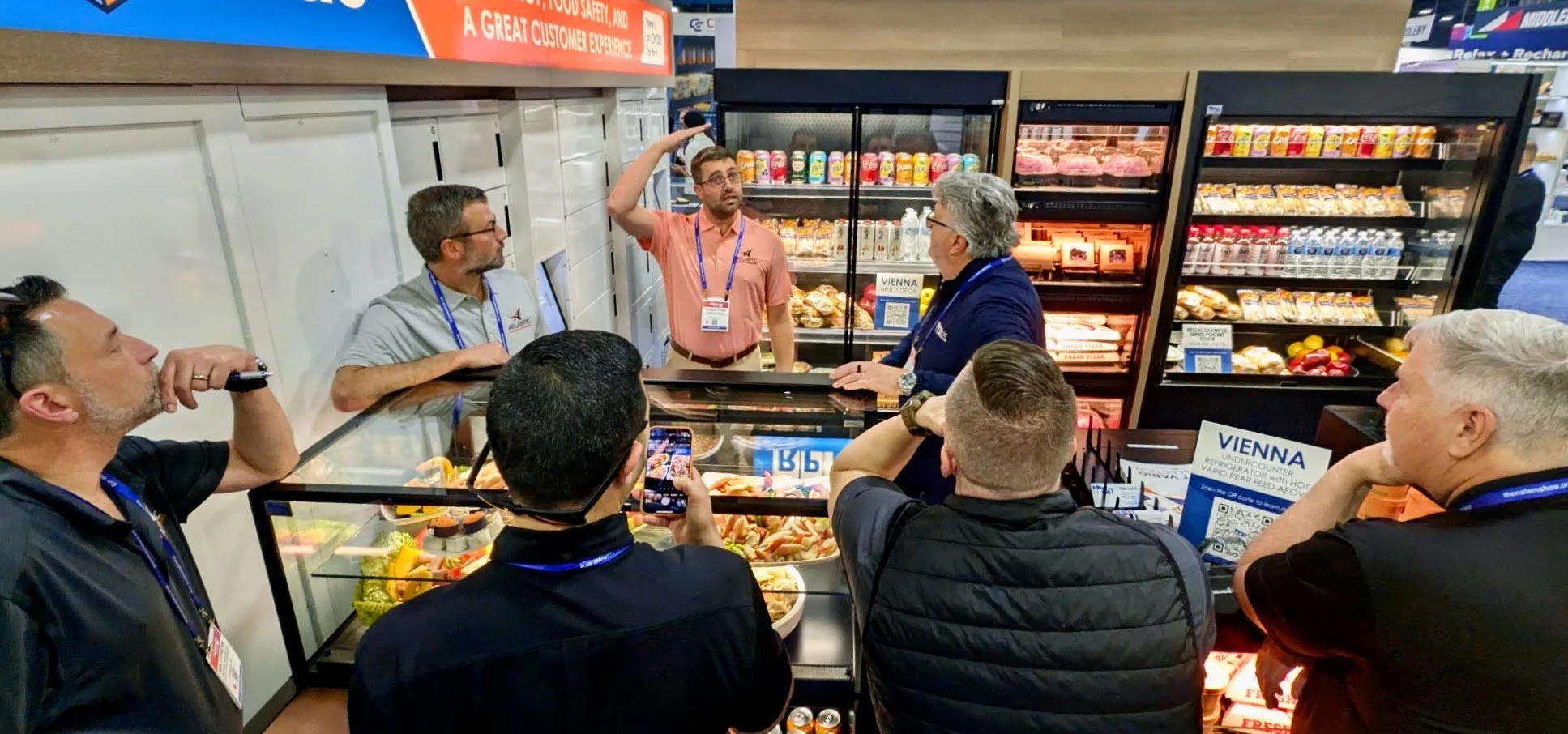 Group of people at an indoor booth, with one person speaking and others listening, surrounded by refrigerated display cases of assorted food items and beverages.
