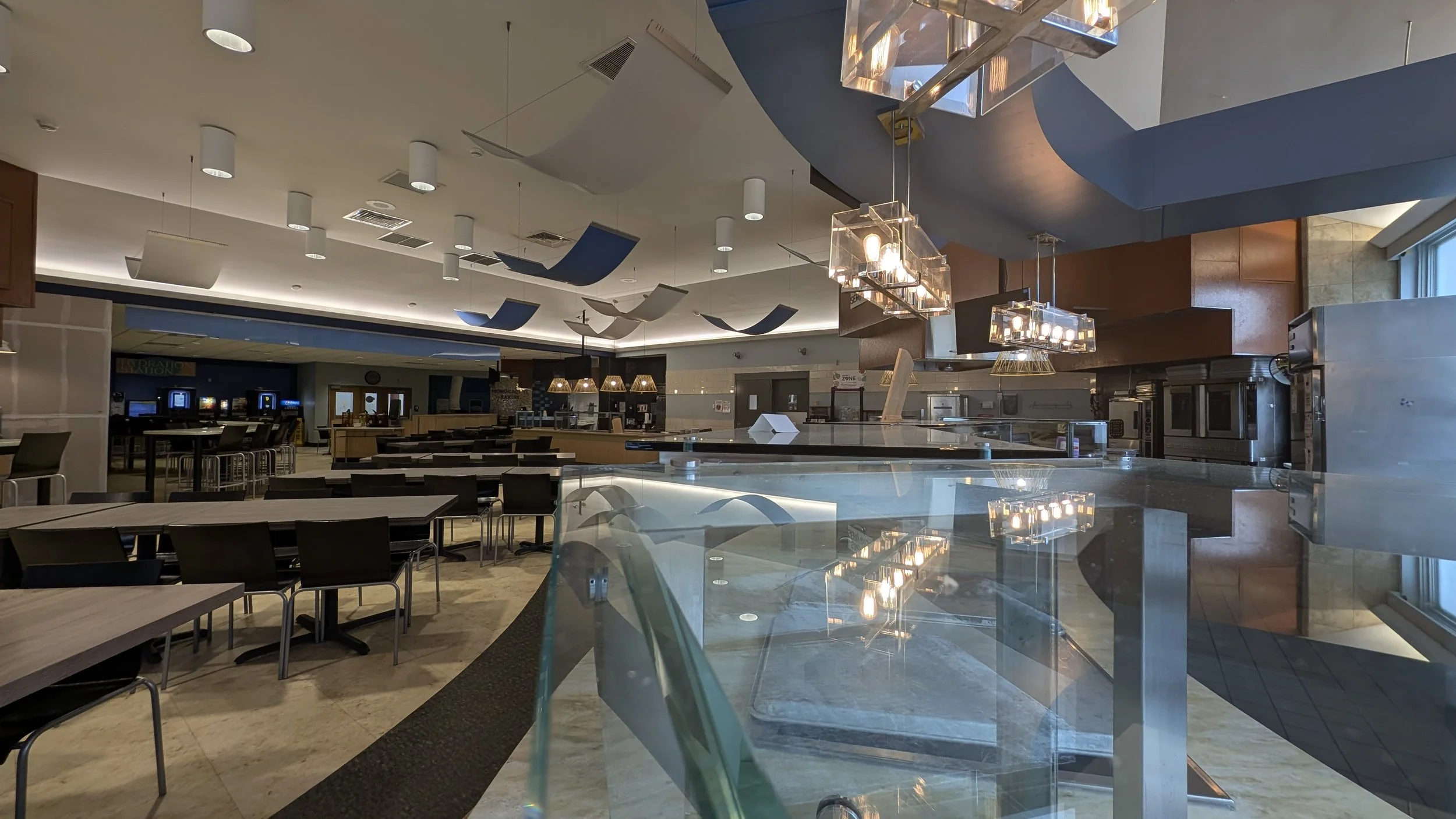 Empty indoor dining area with tables and chairs, modern light fixtures, and ceiling decorative panels in a restaurant or cafeteria.