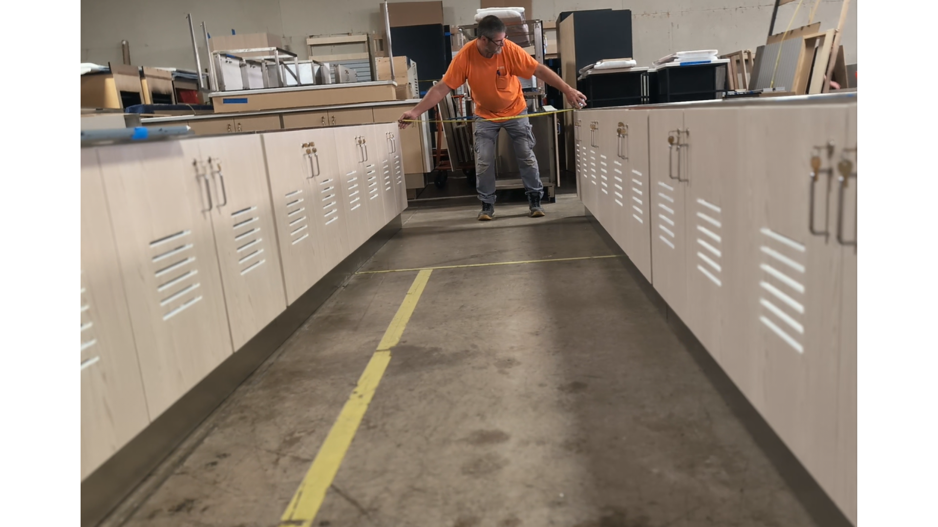 A man in an orange shirt and gray pants measures a cabinet in a workshop with black countertops using a tape measure, standing between two rows of light-colored cabinets.