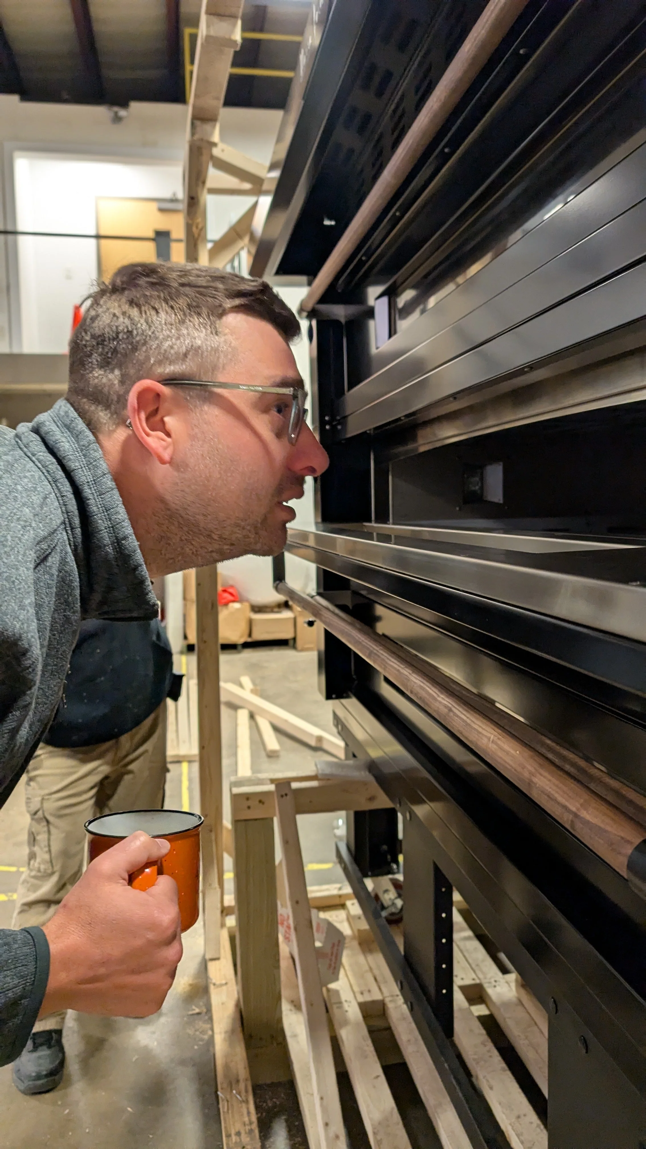 A man with glasses and a gray sweater holds an orange mug and looks closely at a large, black and wood-colored display or equipment inside a workshop or industrial space.