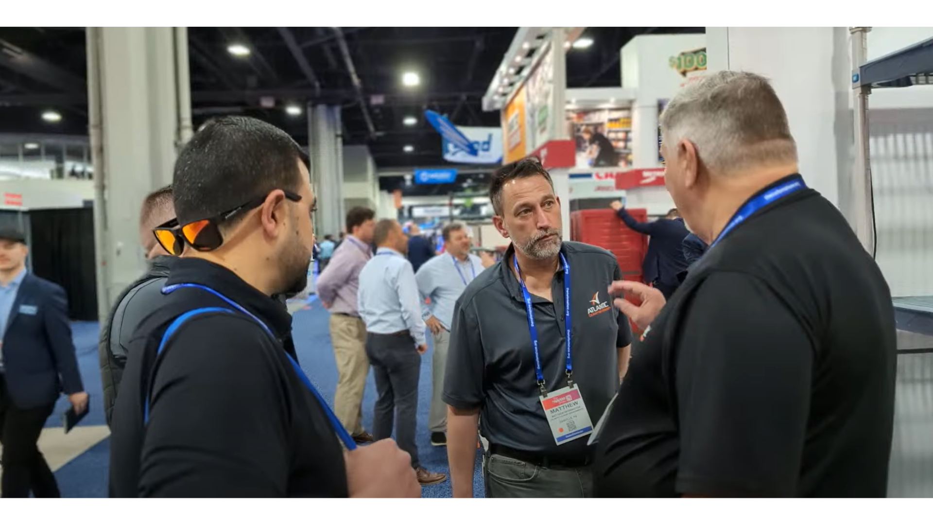 Three men at a trade show or conference engaging in conversation with others in the background.
