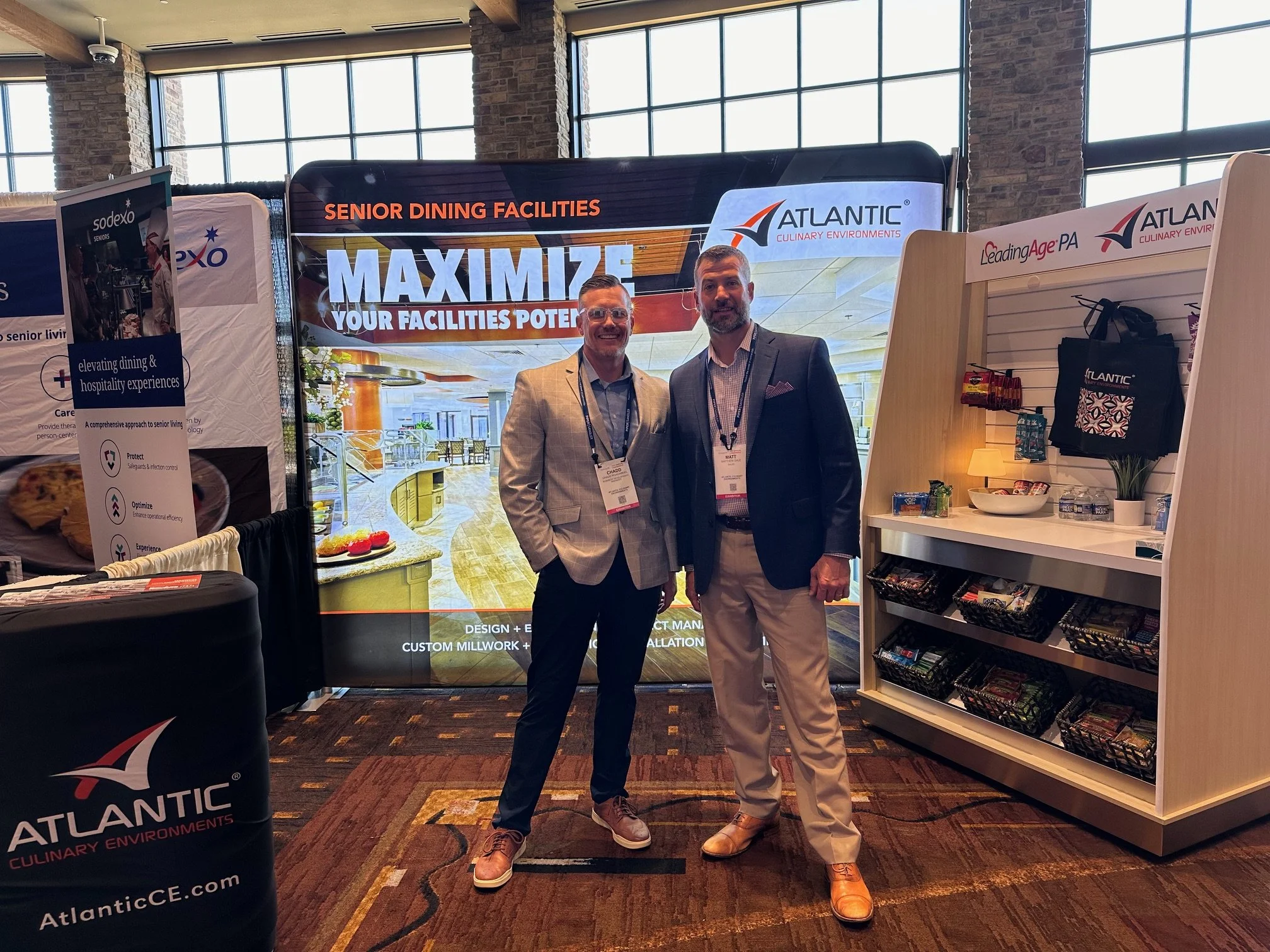 Two men in business attire standing and smiling at a conference booth with signage reading 'Maximize Your Facilities Potential' and 'Atlantic Culinary Environments' in a large, well-lit room with windows and brick walls.