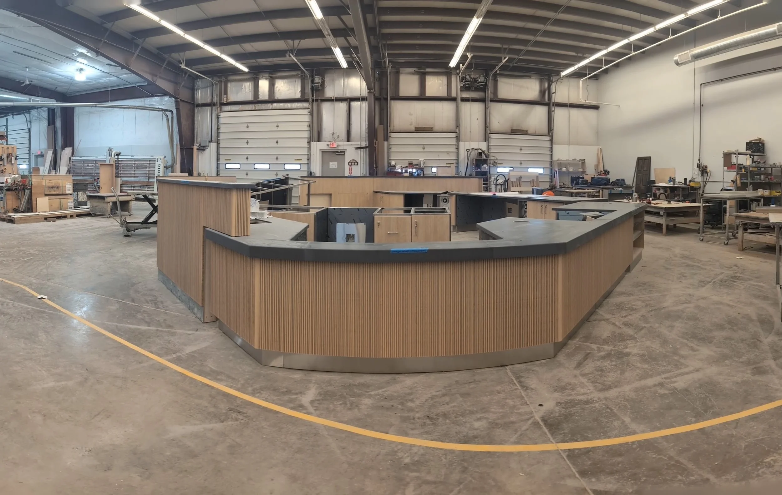 An empty workshop or fabrication shop with a curved wooden reception desk in the center and various tools and materials on workbenches along the walls.