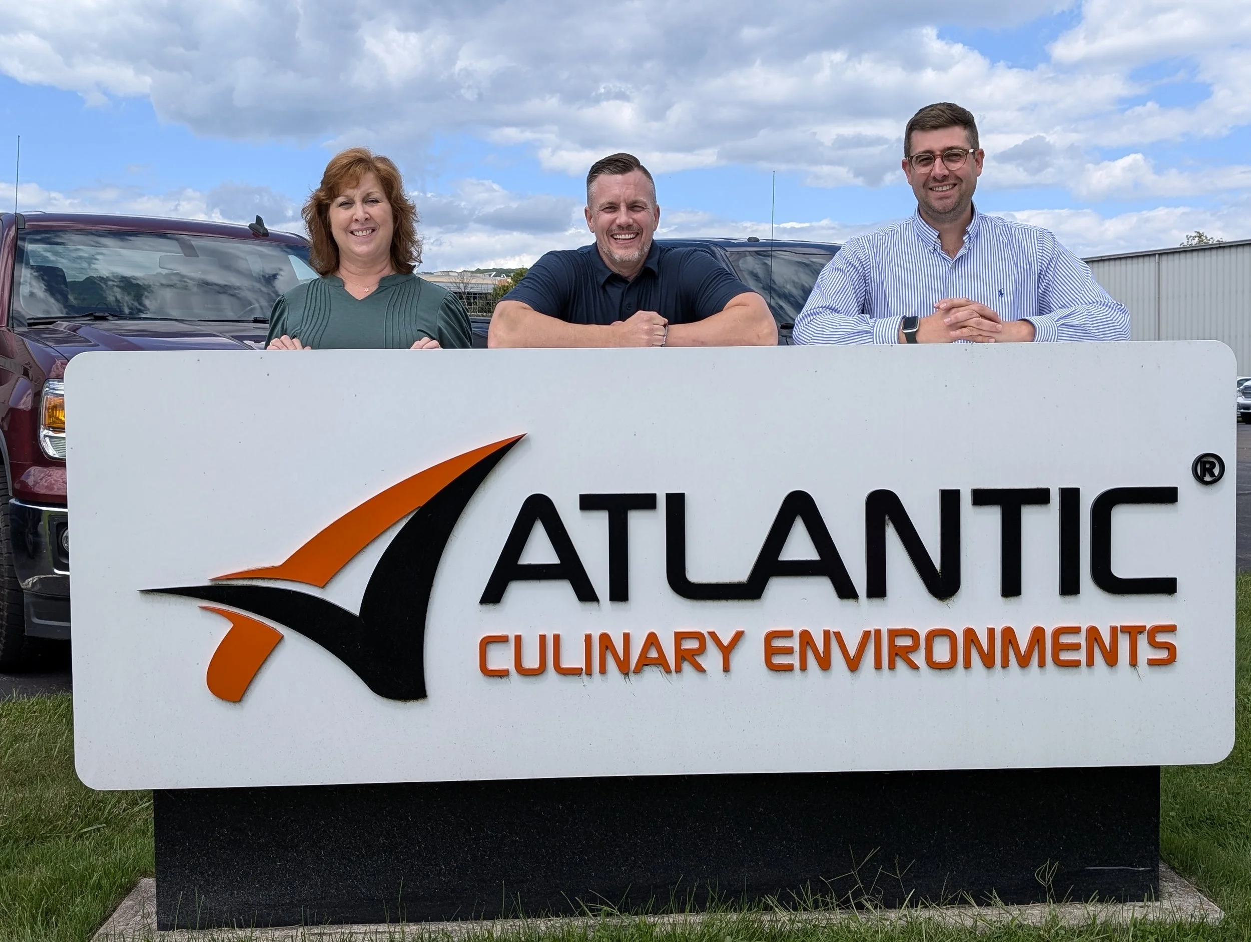Three people standing behind a sign that reads "Atlantic Culinary Environments" with a stylized logo, outdoor setting, cloudy sky above.