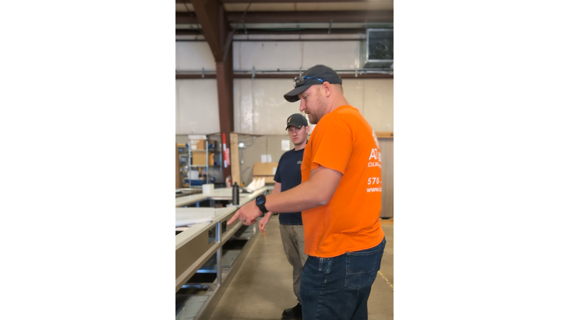 Two men in a warehouse looking at a long work table with equipment. The man in the foreground is wearing an orange shirt, a black cap, and a smartwatch, and is pointing at something on the table. The man in the background is wearing a black shirt and