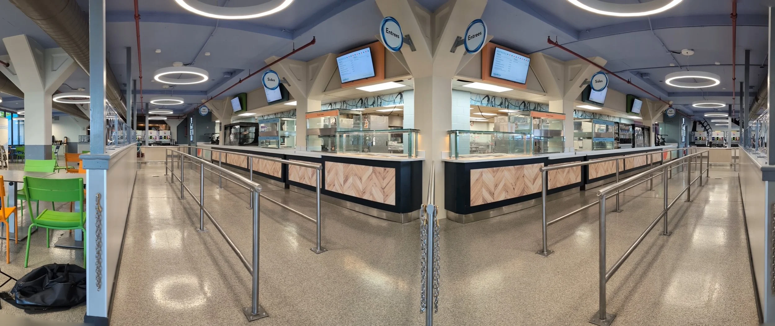 Empty cafeteria food service area with colorful chairs, counter orders labeled 'Entrees' and 'Sides', digital menu screens, and metal railings.