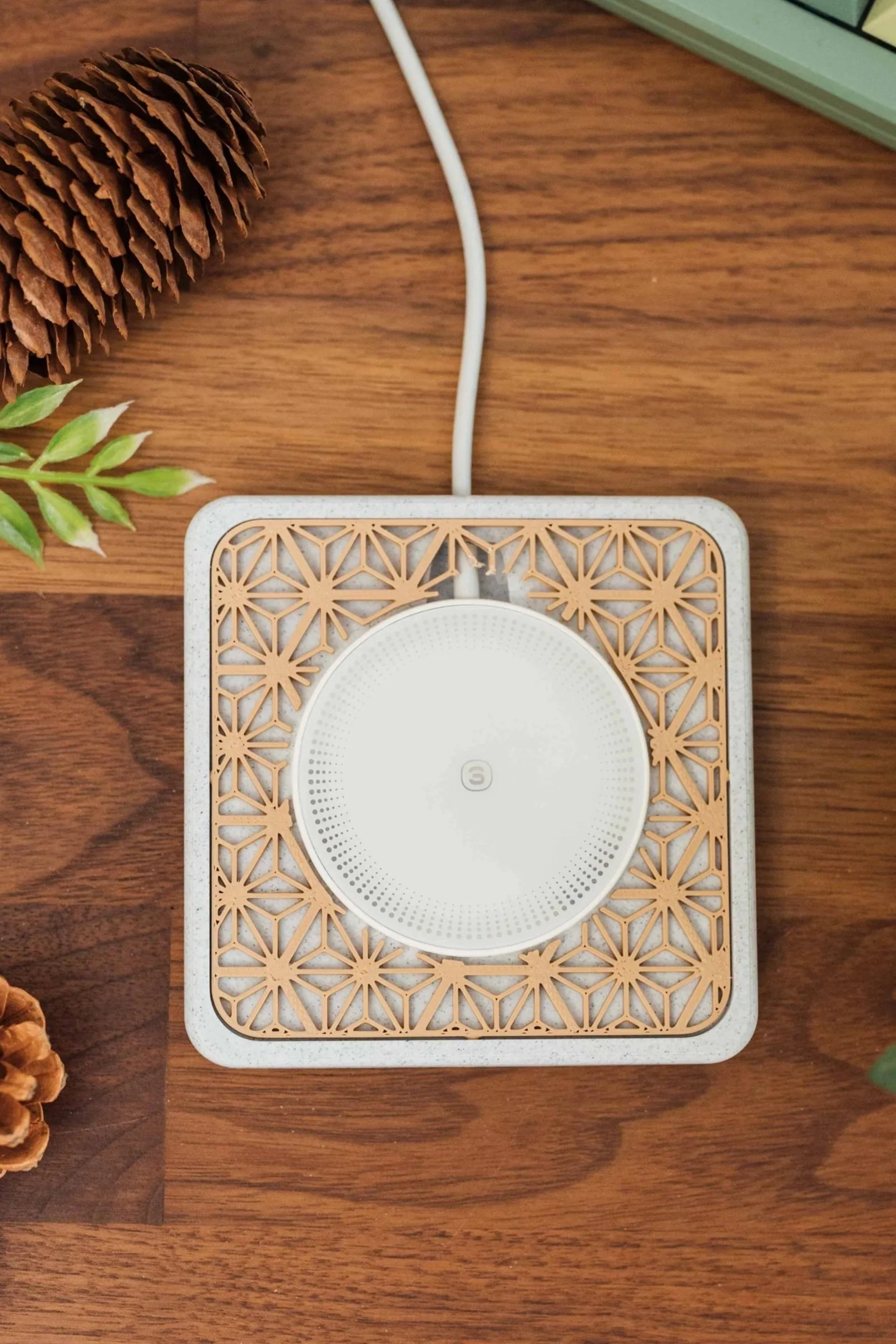 A white smart speaker placed on a decorative wooden grid, on a wooden surface, with plants and pinecones around it.
