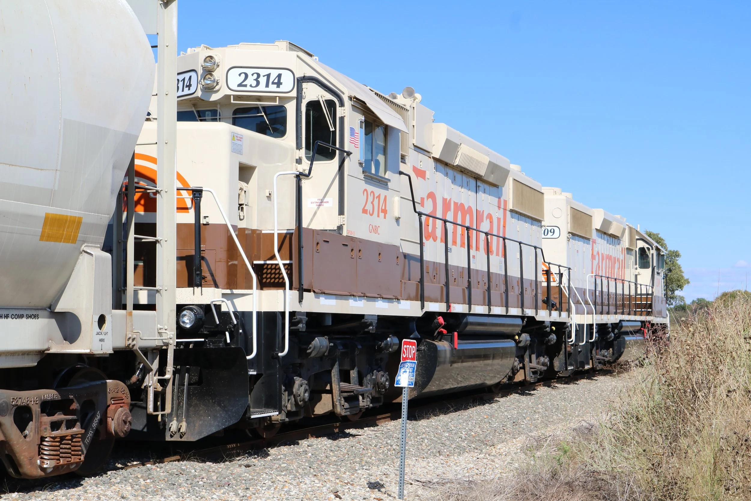 Two freight trains sitting on railroad tracks in a rural area with clear blue sky. One is a white and brown locomotive with the number 2314, and the other is a white locomotive in the background.