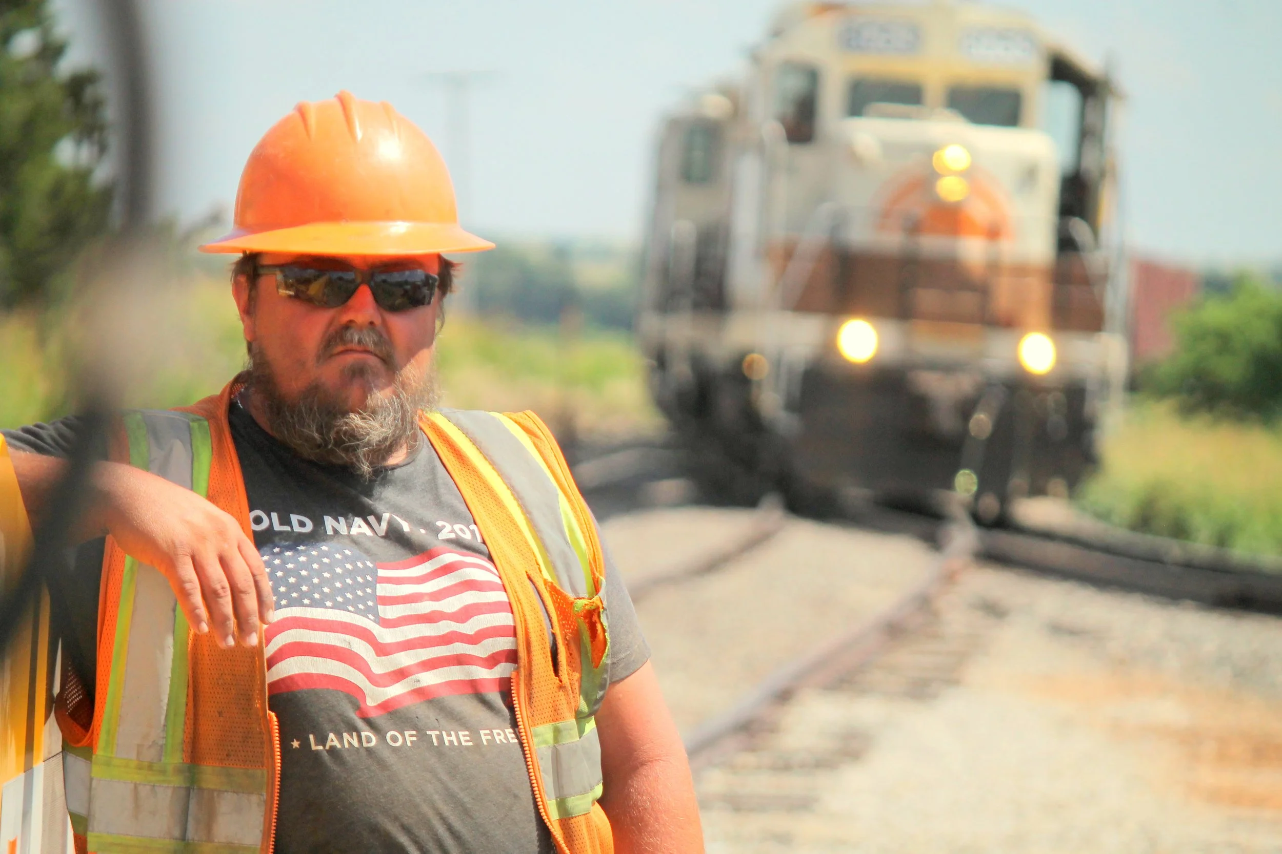 A man wearing an orange hard hat, sunglasses, and a reflective safety vest, posing with his hand on his hip in front of a moving train on a railway track.