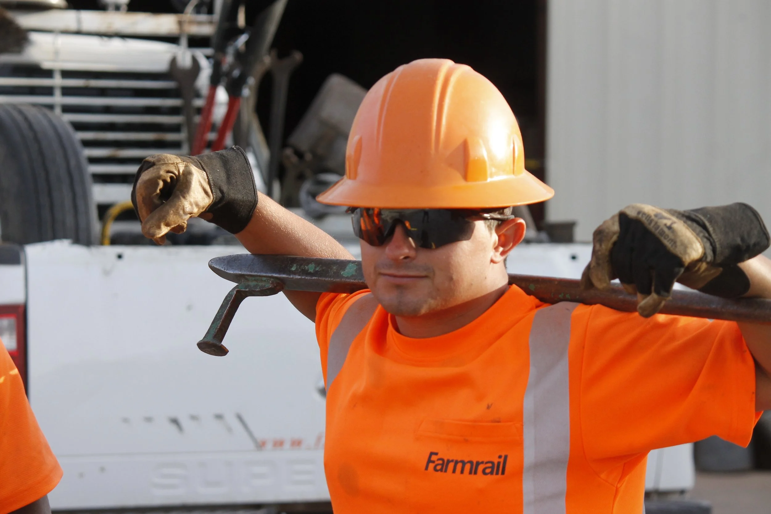A construction worker wearing an orange safety shirt, a hard hat, and sunglasses, carrying a shovel over his shoulders.