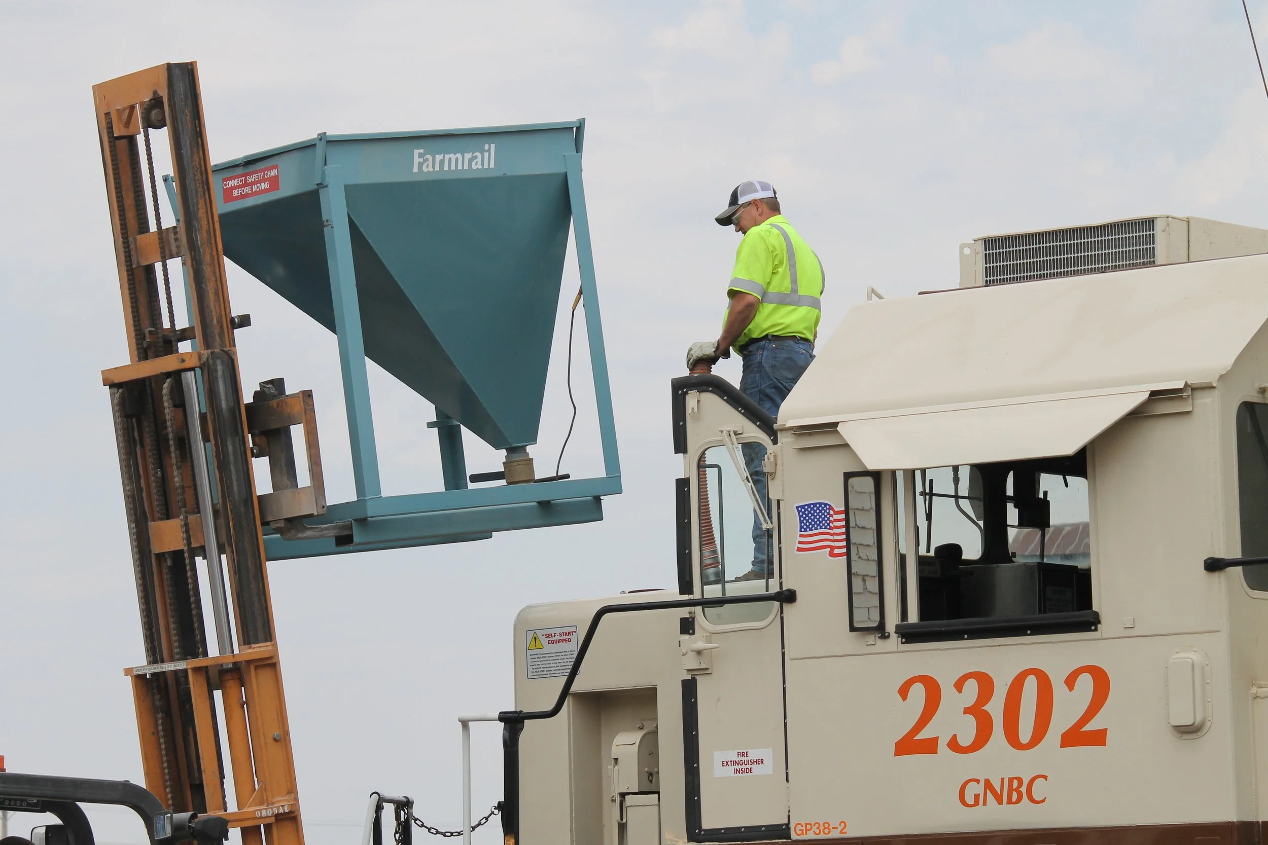 A worker in a yellow safety vest and cap is standing on an elevated platform of a construction vehicle, working on a blue machine labeled 'Farrmal' against a cloudy sky.