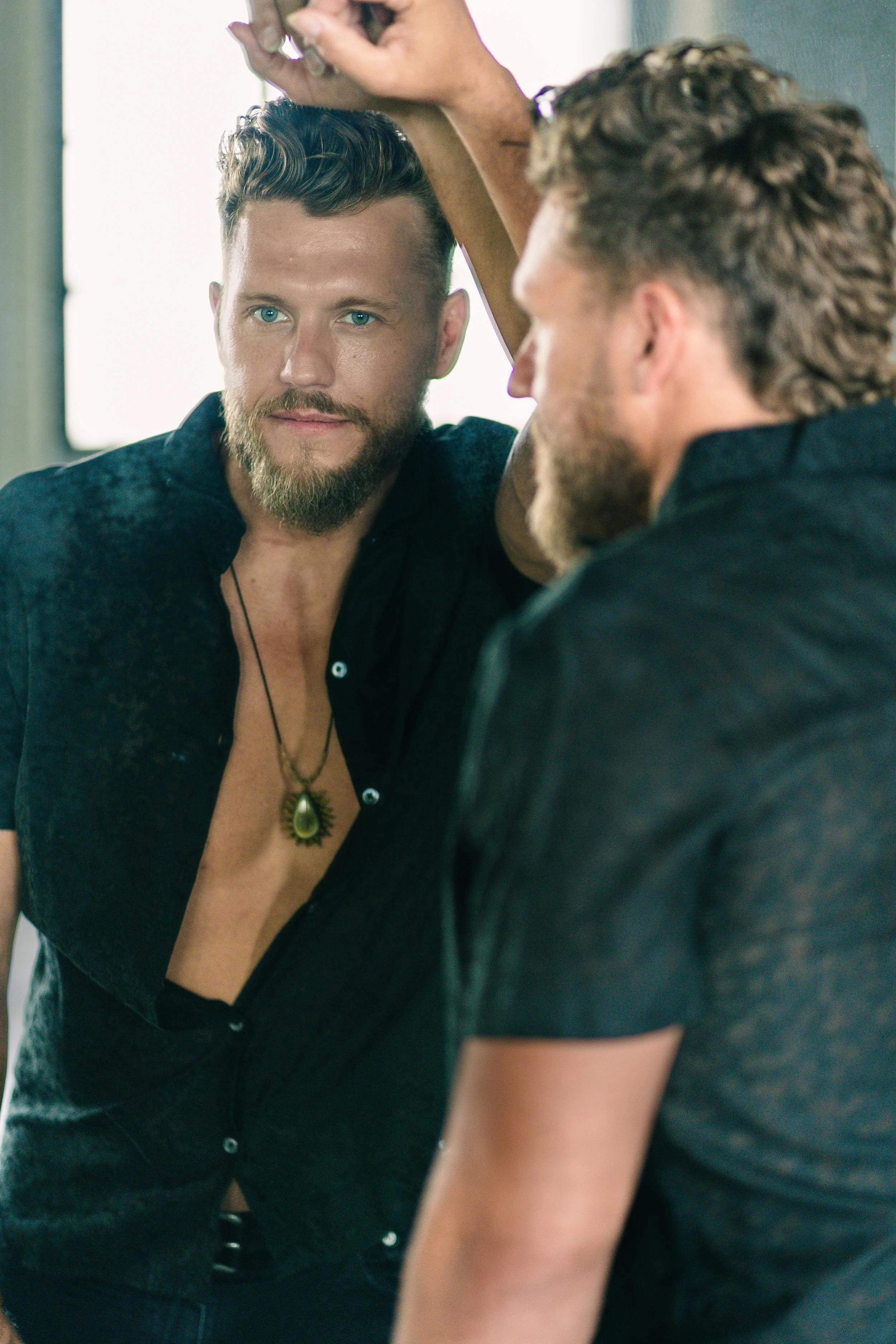 A man with a beard and styled hair looking into a mirror, wearing an open black shirt and a pendant necklace, with his reflection visible.