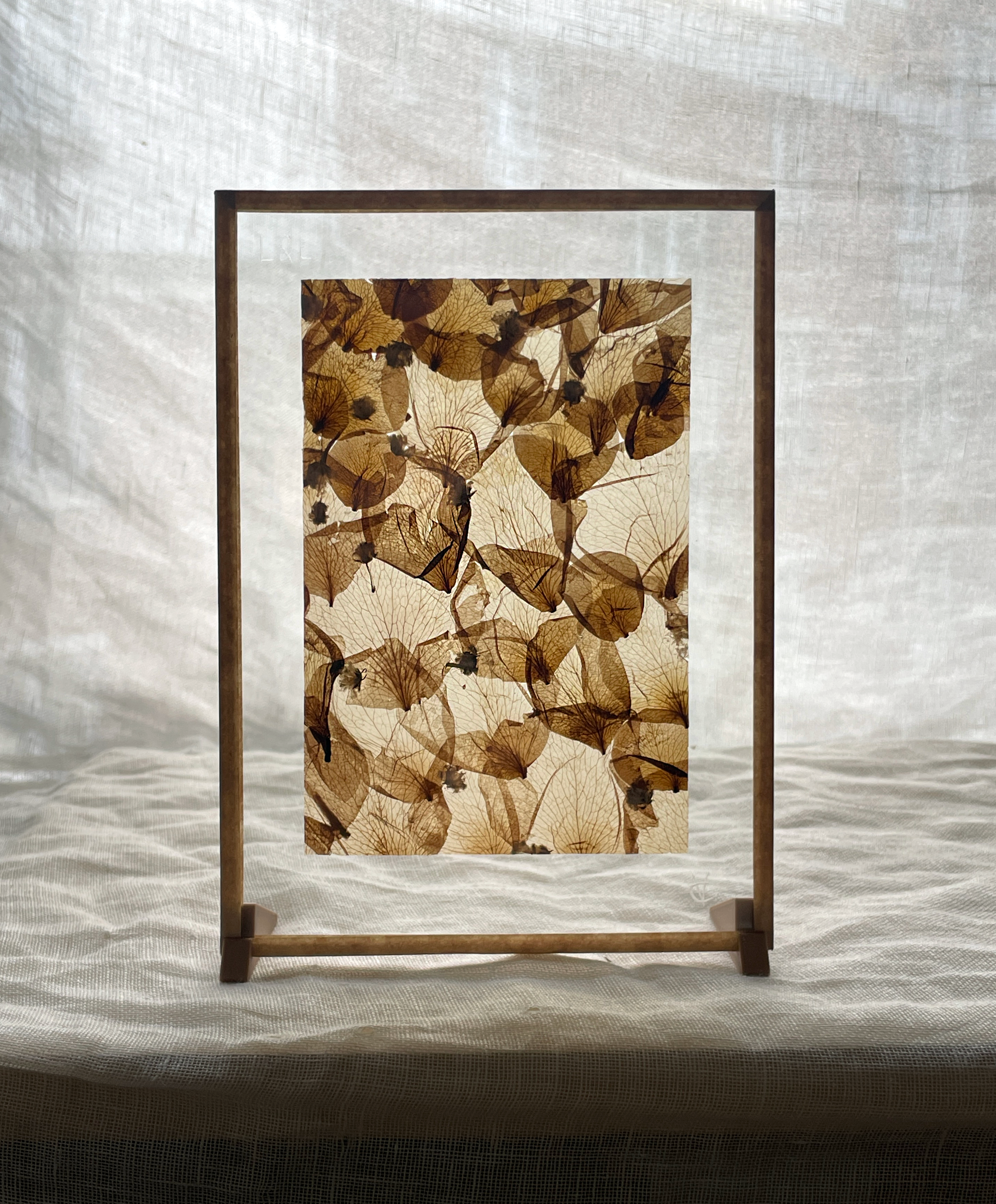 Cadre en bois avec une feuille de papier à motif de feuilles d'automne, posé sur une table en tissu, devant un rideau blanc.
