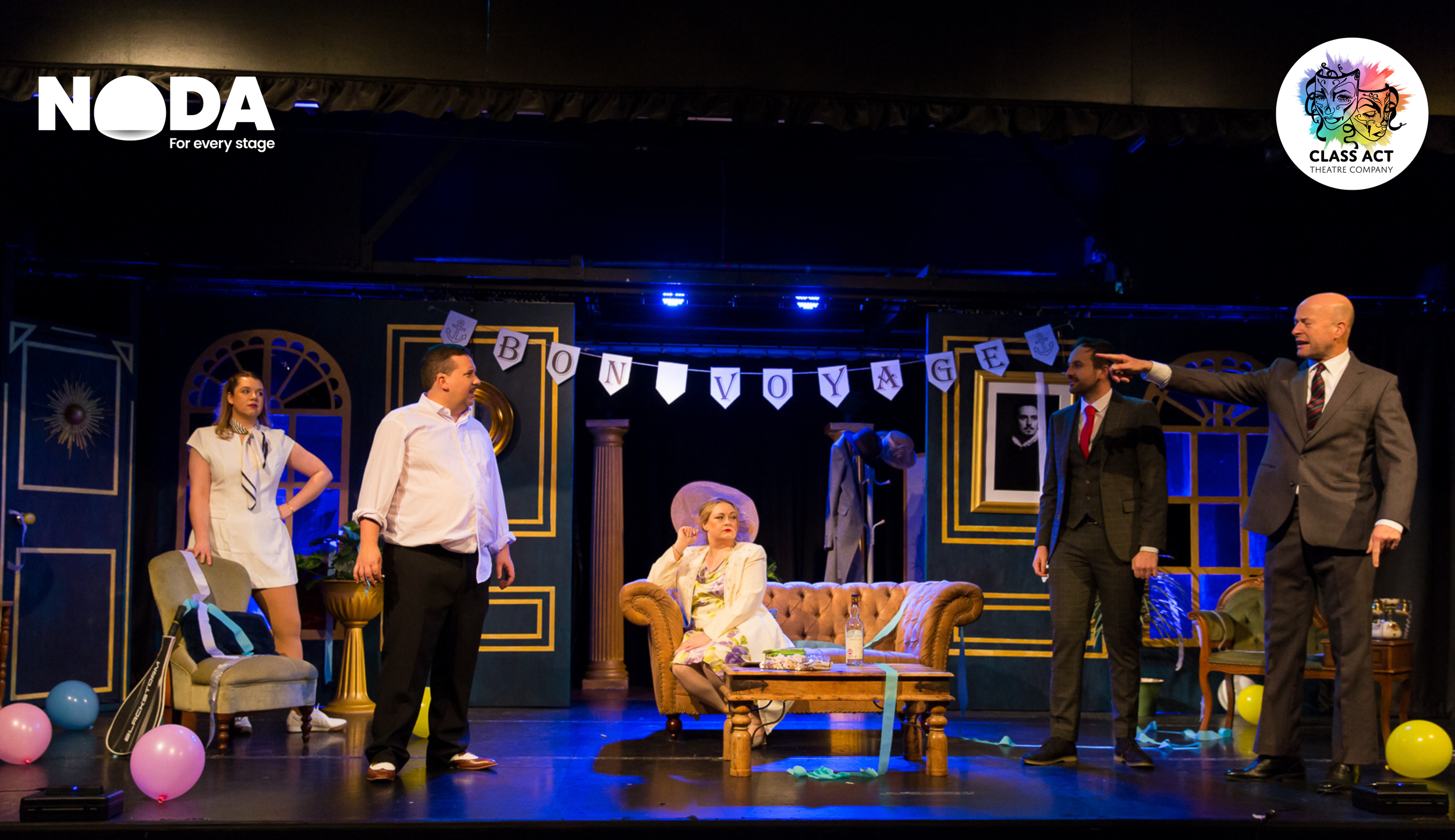Theater stage scene with five actors, decorated with balloons and a banner that reads 'BON VOYAGE'. An older woman is seated on a sofa holding a large hat, while the others are standing around her, engaged in conversation. The set features framed pictures, columns, and a framed portrait, creating an elegant, vintage atmosphere.