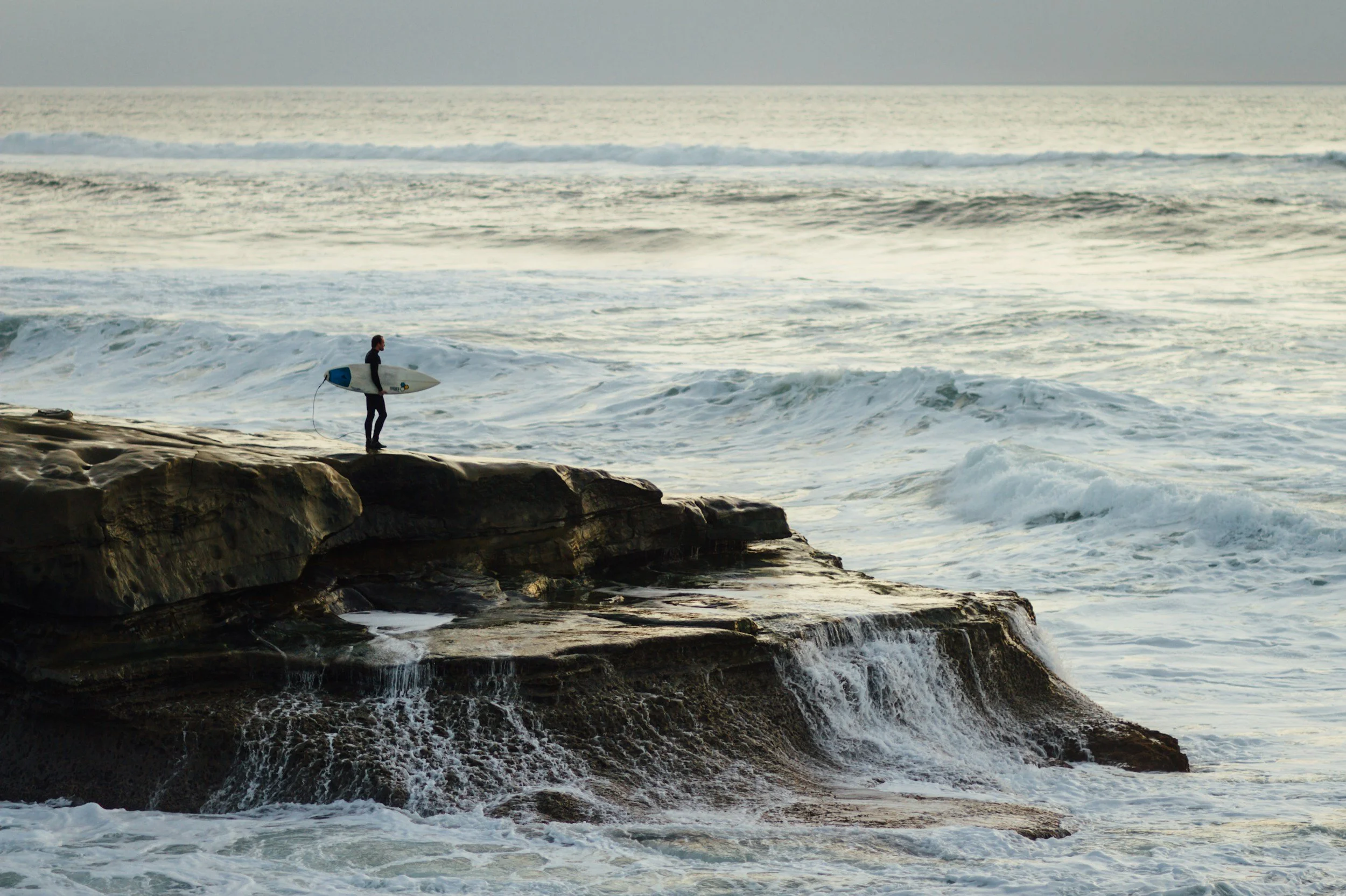A person standing on rocks at the beach, holding a surfboard, looking at the ocean waves and horizon during a cloudy day.