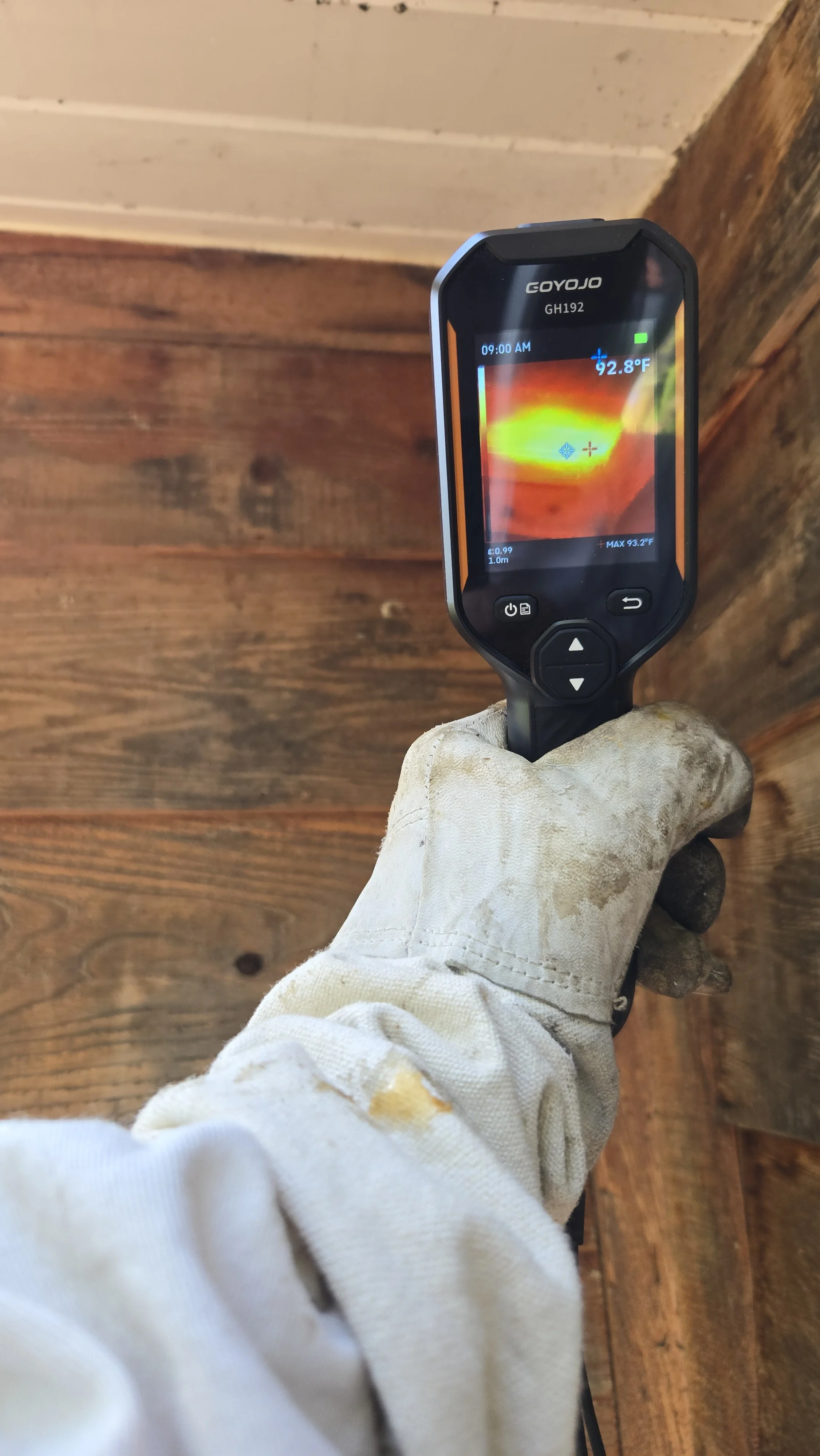 A person's hand wearing a white work glove holding a COYOJO infrared thermometer pointed at a wooden surface. The thermometer display shows a temperature of 92.8°F and a time of 9:00 AM.