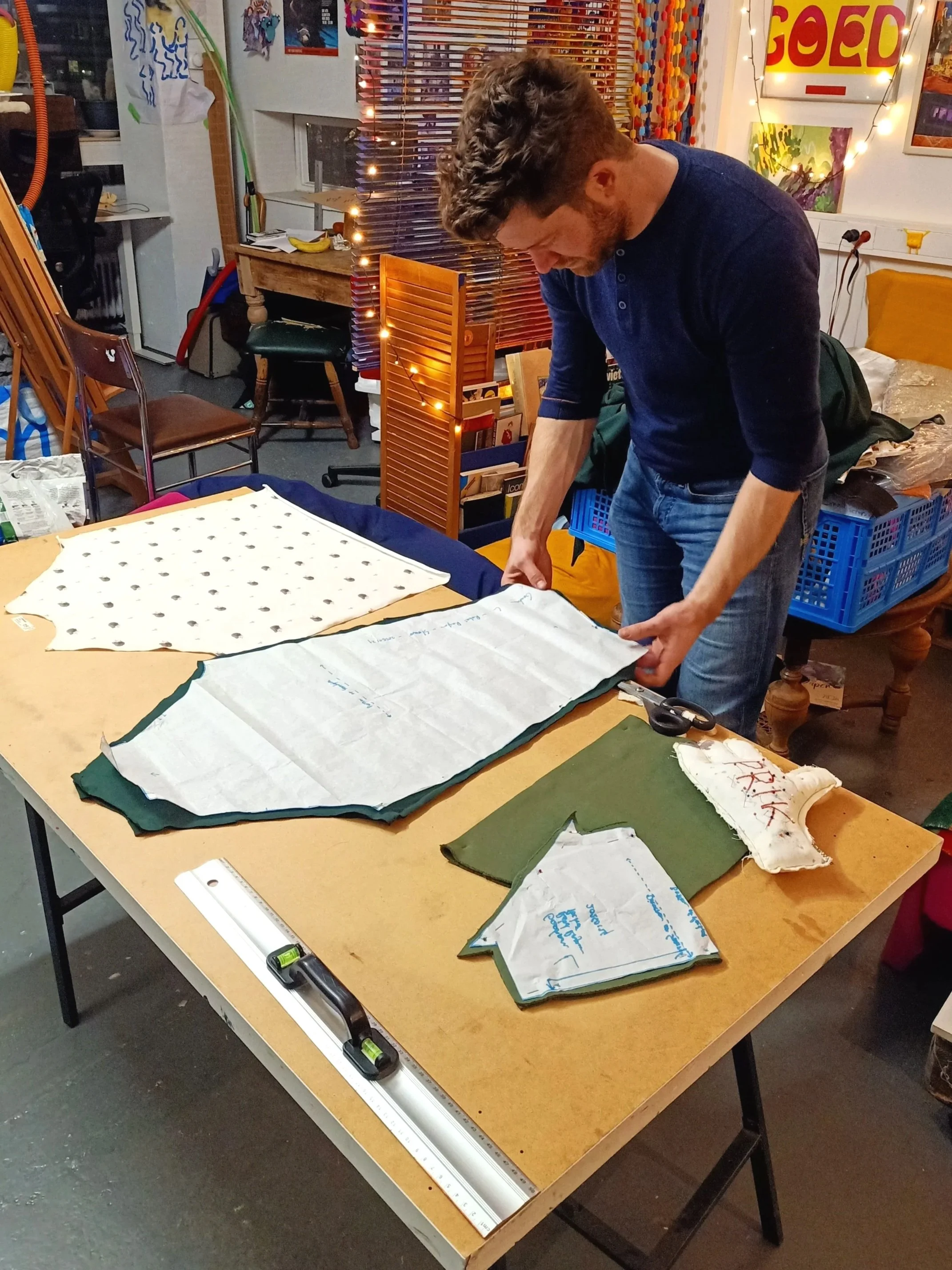 Man die stoffen snijdt op een tafel in een creatieve werkomgeving met lampjes en kleurrijke kunstwerken op de muur.