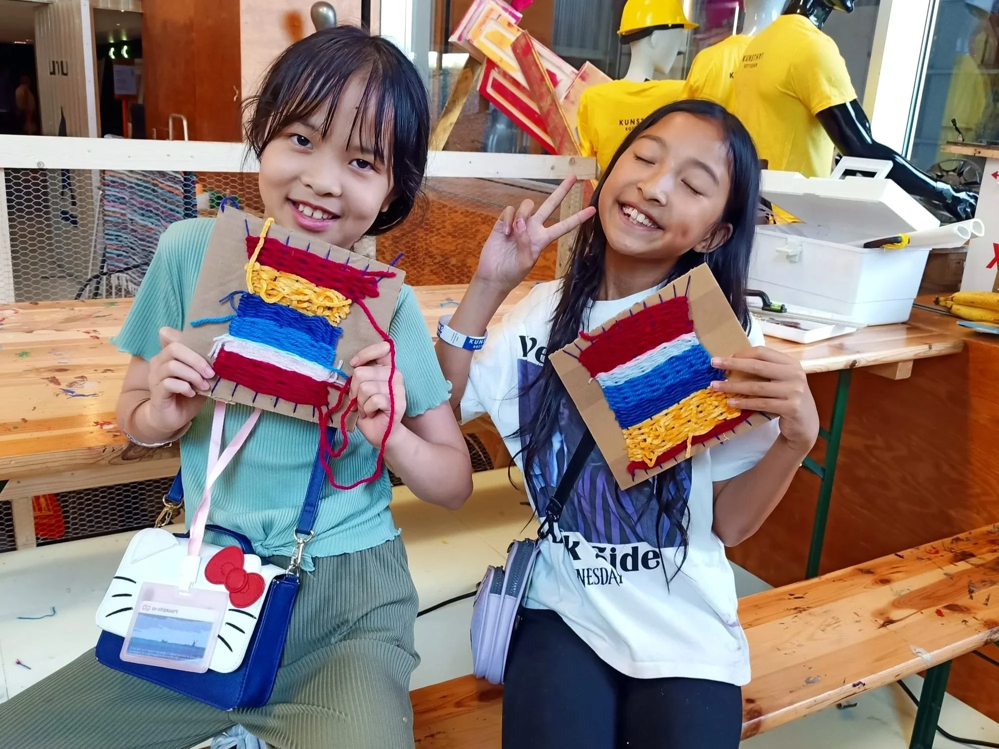 Twee jonge meisjes poseerden met handgemaakte textielkunstwerken in een knutselruimte. Ze lachen en maken vrede-gebaar while holding hun kunstwerken. Op de achtergrond zijn mannequin koppen en t-shirts met teksten te zien.
