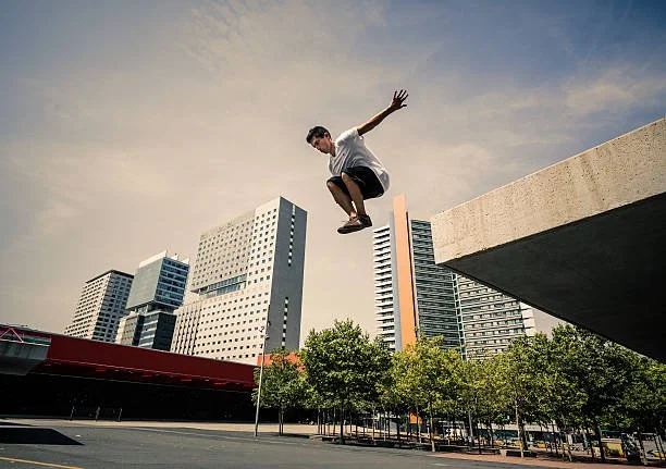 A person in mid-air jumping from a ledge in an urban area with modern high-rise buildings and trees below.