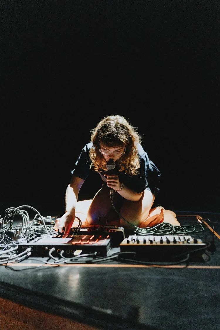 A woman kneeling on a dark stage, surrounded by electronic music equipment, holding a microphone and adjusting a gadget.