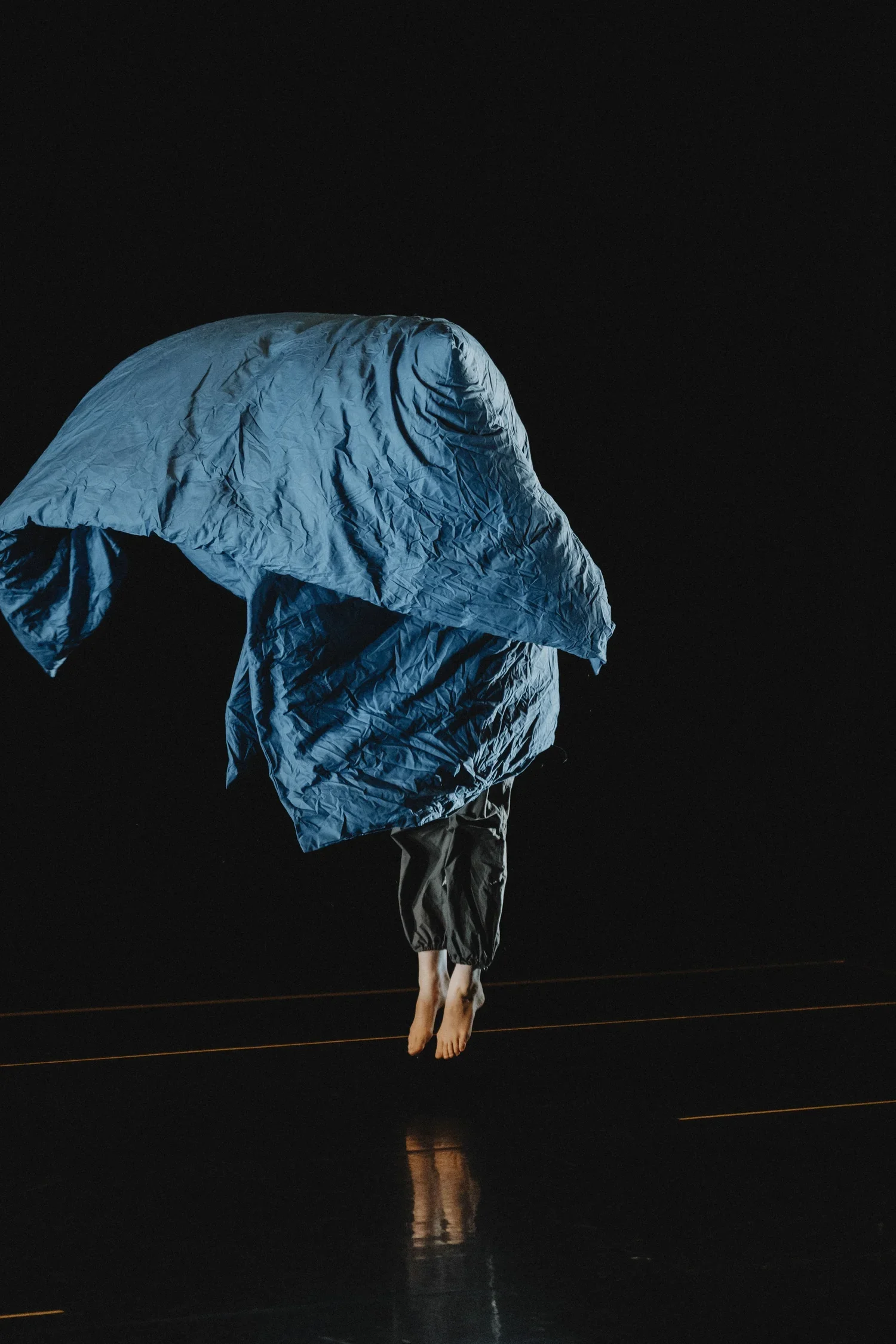 Person hanging upside down with a large blue fabric or parachute covering their head, barefoot, against a black background.