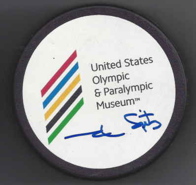 A round badge with the United States Olympic & Paralympic Museum logo and signature, featuring colorful stripes and black, red, yellow, blue, and green lines.