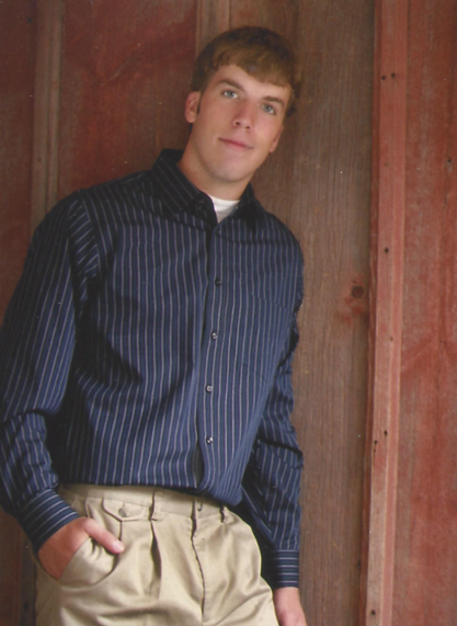 A young man with light brown hair wearing a navy blue striped dress shirt and beige pants, standing against a wooden wall.