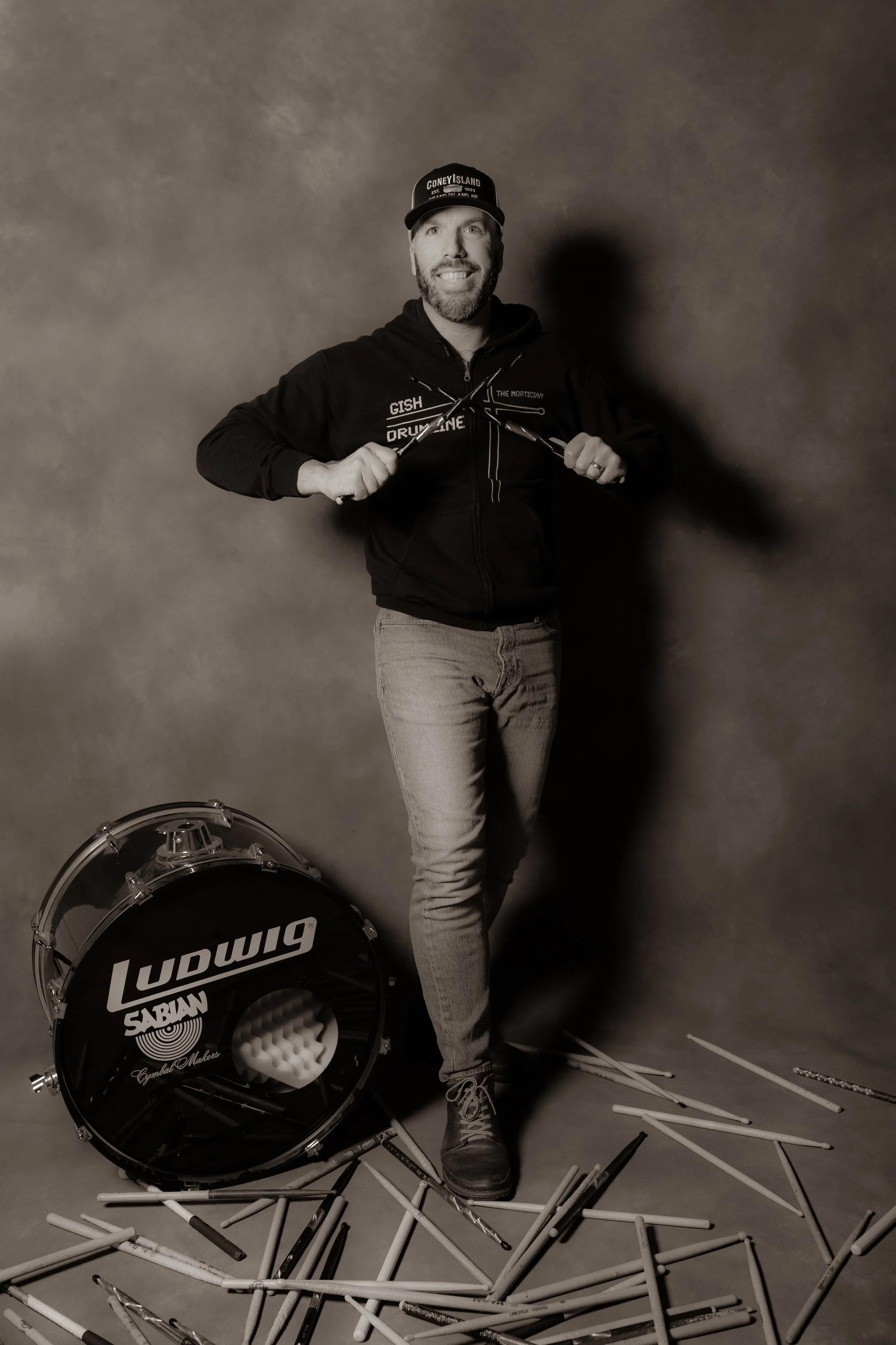 A man with a beard and a hat standing amidst scattered drumsticks, holding two drumsticks crossed in front of his chest, with a Ludwig bass drum at his feet, in a photography studio setting.