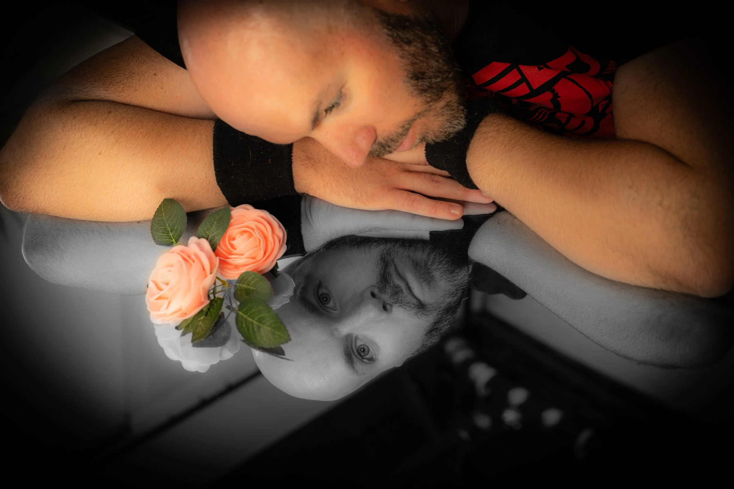 Man resting his head and arms on a surface, with a black and red shirt, close to a mannequin head with painted facial features, wearing a pink rose flower crown with green leaves, against a black and white background.