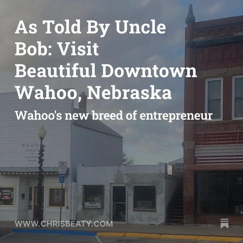 A street view of downtown Wahoo, Nebraska, with a white building on the left and a red brick building on the right, under a cloudy sky.