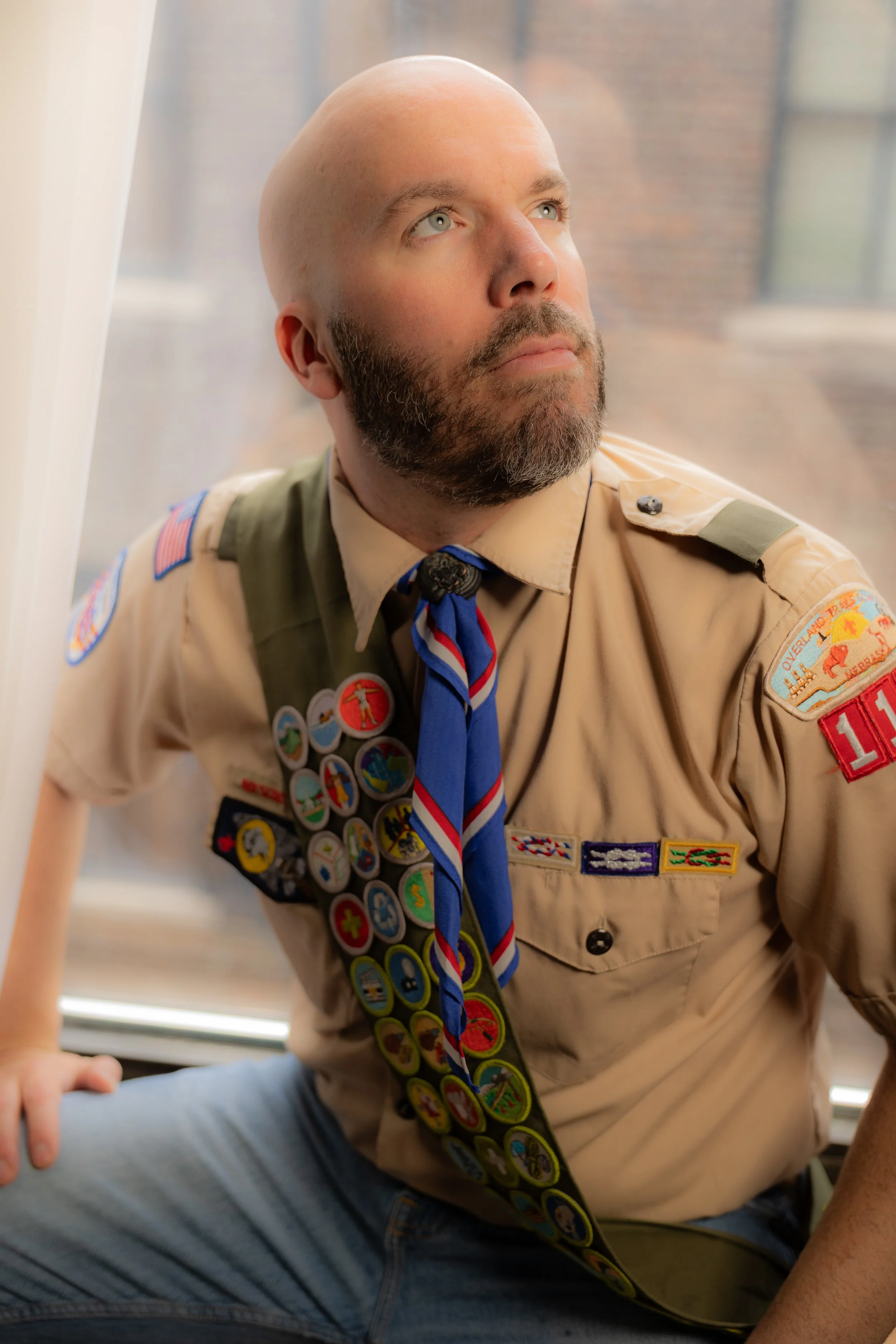 A man in a Cub Scout uniform with scouting badges and a neckerchief, sitting by a window and looking upward.