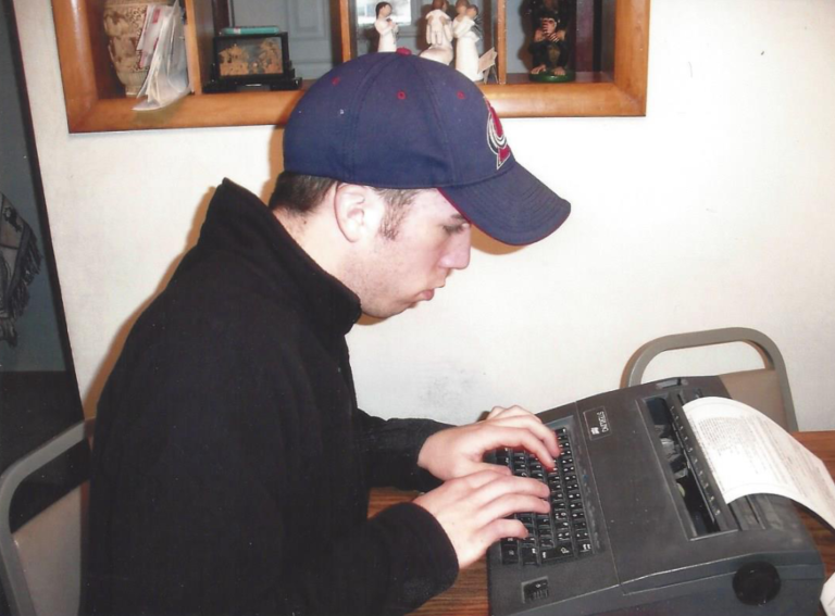 A young man in a dark hoodie and cap typing on a vintage typewriter, with a closed book nearby, in a room with wooden framed mirror and decorative dolls on a shelf.