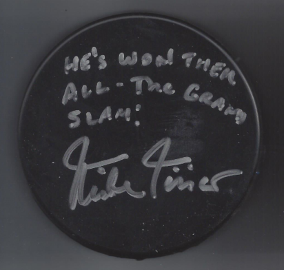 Inscription on a black hockey puck reading, "He's word tinker all--the cream slap!" with a signature below.
