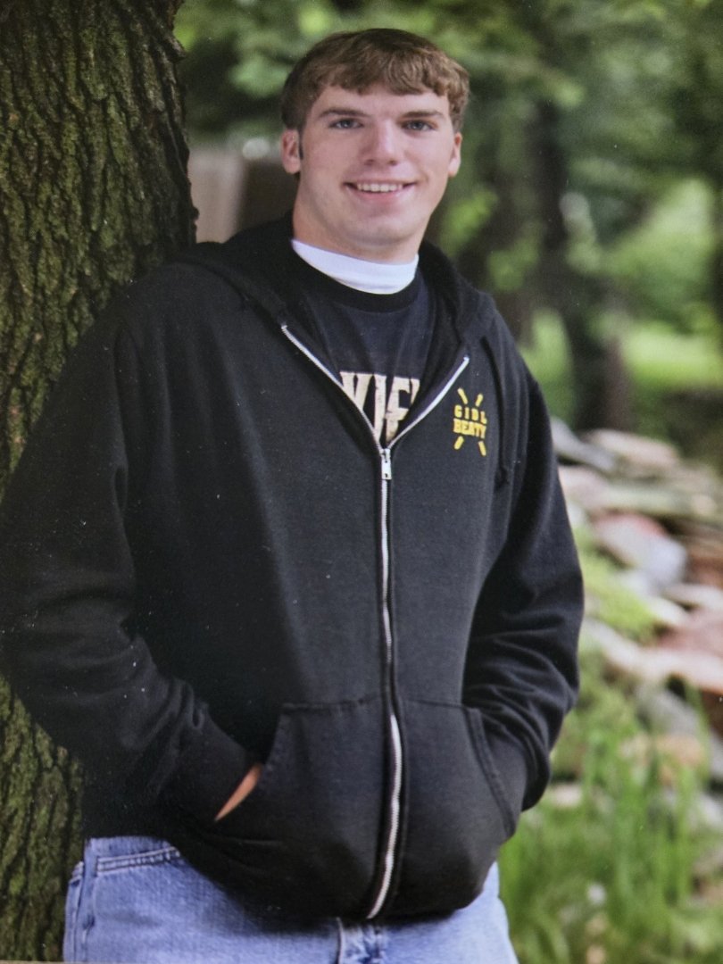 A young man with brown hair wearing a black hoodie with yellow graphics standing outdoors near a tree with lush green foliage in the background.
