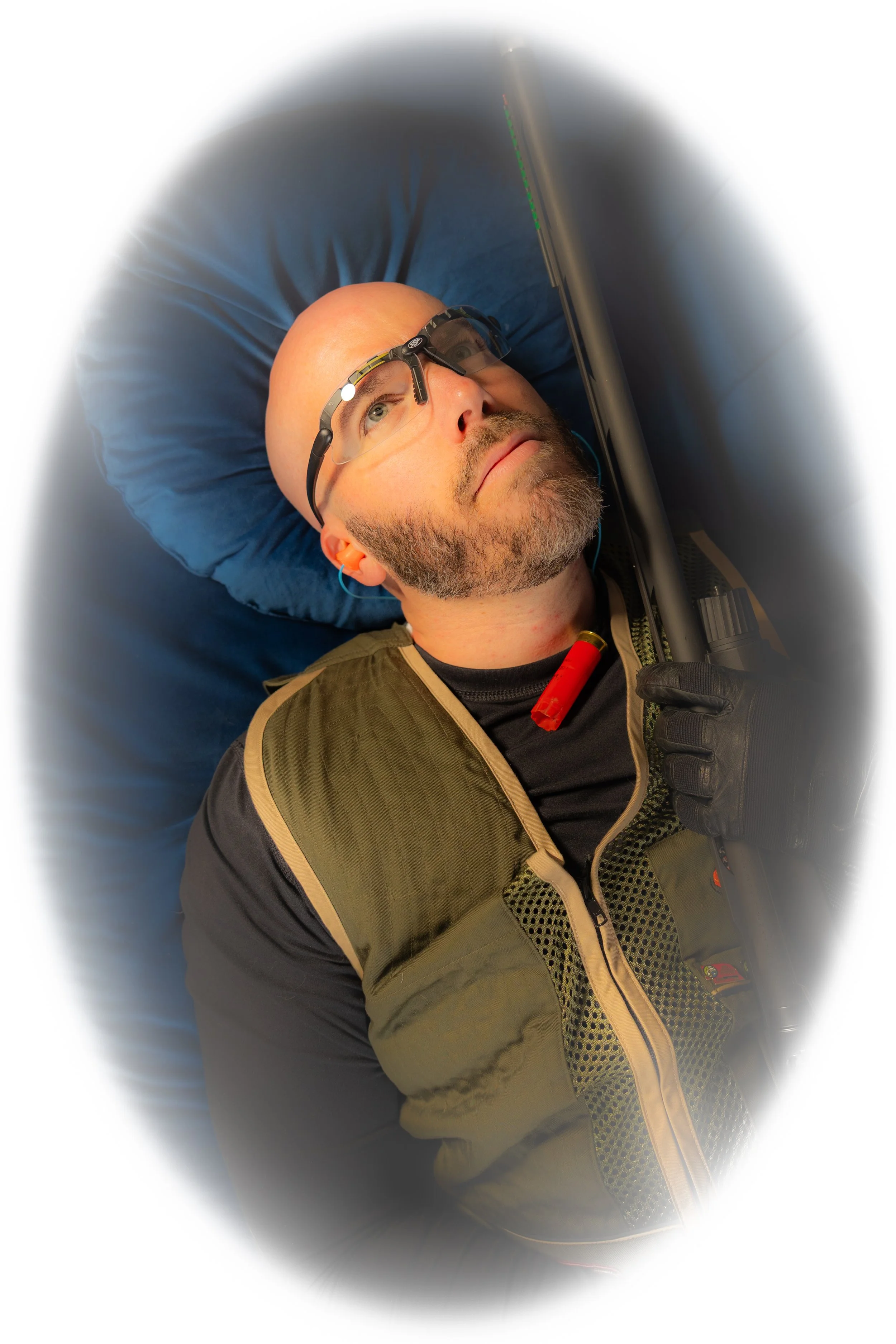A man lying on a blue pillow, looking upward, wearing protection glasses, holding a firearm with gloved hands, in an indoor setting.