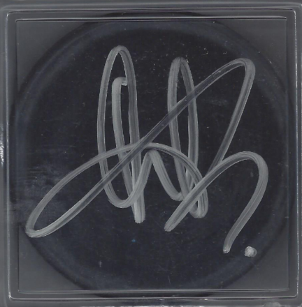 A black circular surface with a silver autograph signature and a dot, inside a clear plastic frame.