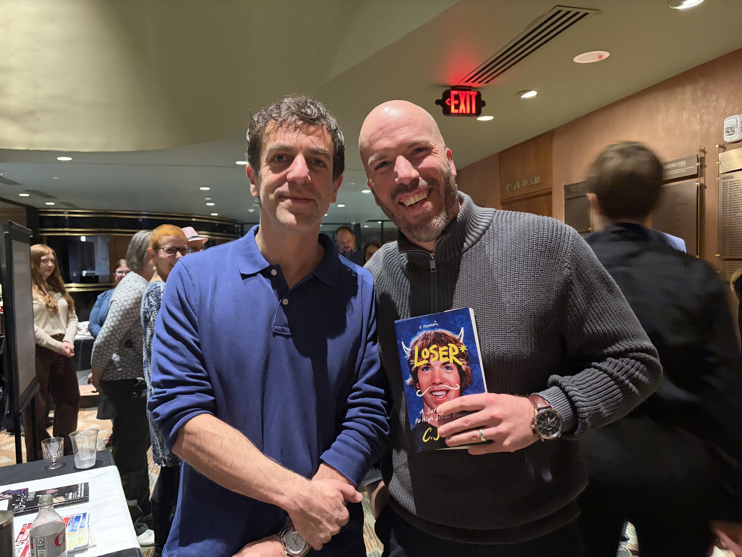 Two men smiling and shaking hands at an indoor event, one holding a book titled 'LOSER' with a cartoon face on the cover, surrounded by other people in the background.