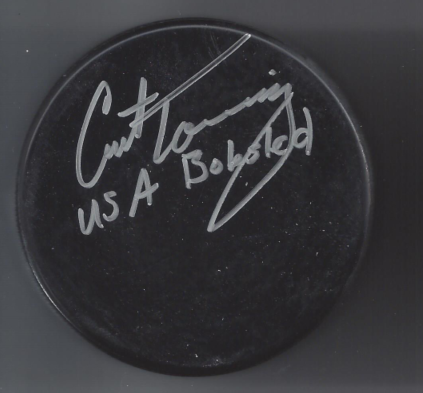 A black hockey puck with a white signature and the words 'U.S.A. BOBBED' written on it.