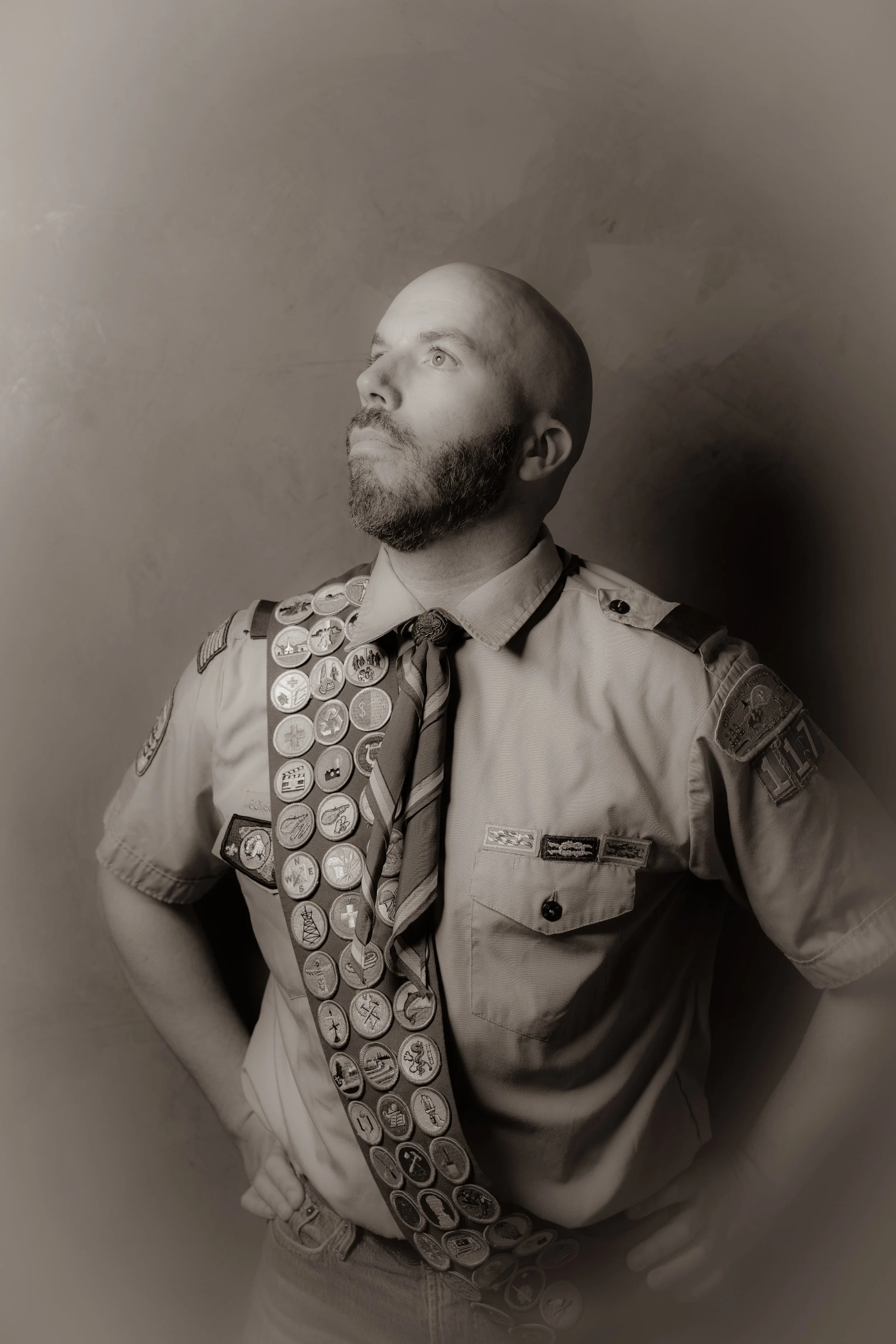 A man in Boy Scout uniform with numerous merit badges on his sash, standing confidently with his hands on his hips, looking upwards.