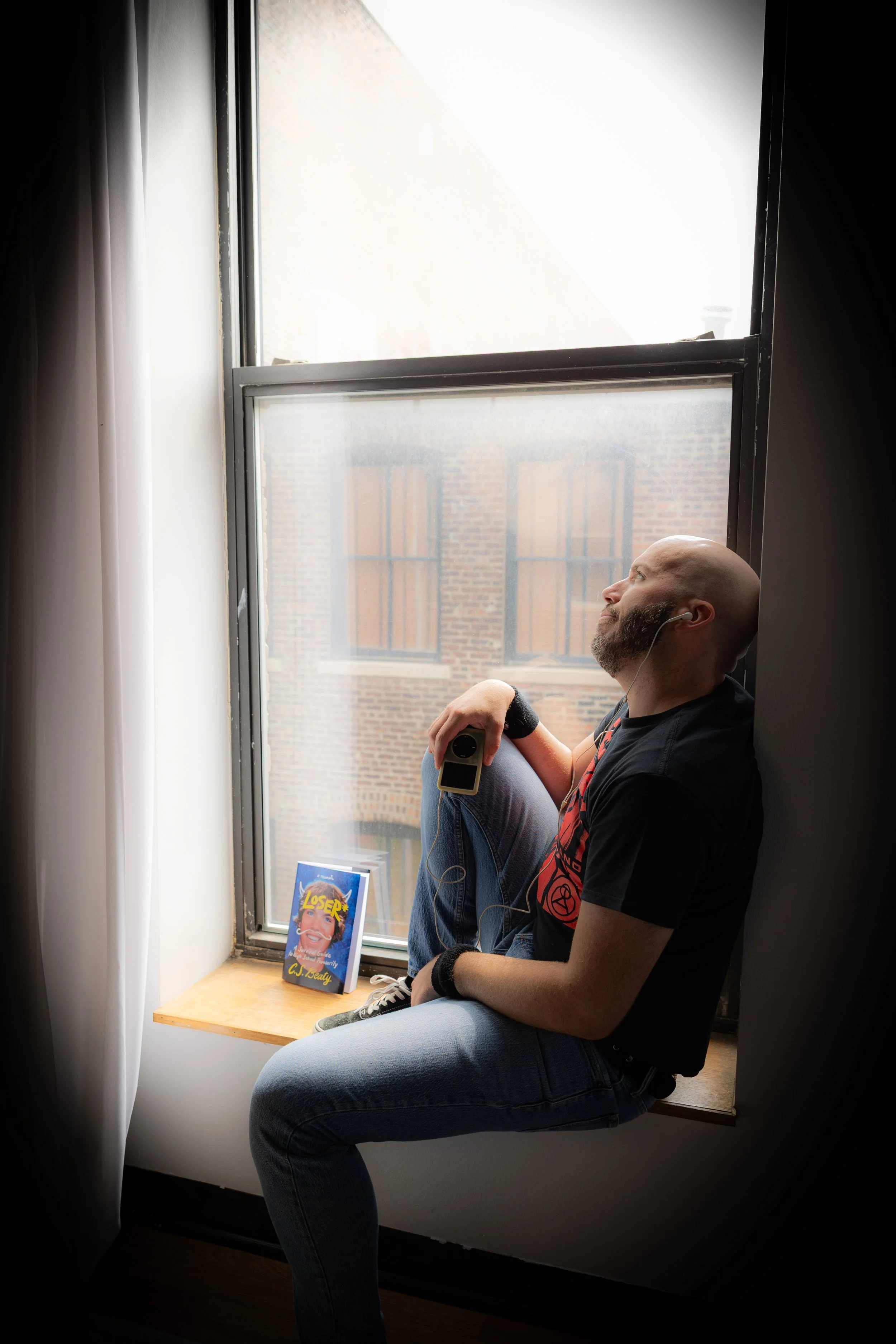 A man sitting by a window, listening to music with earbuds, holding a remote, and looking relaxed. There is a book titled 'LOSER' by CJ Beuaty on the window ledge.