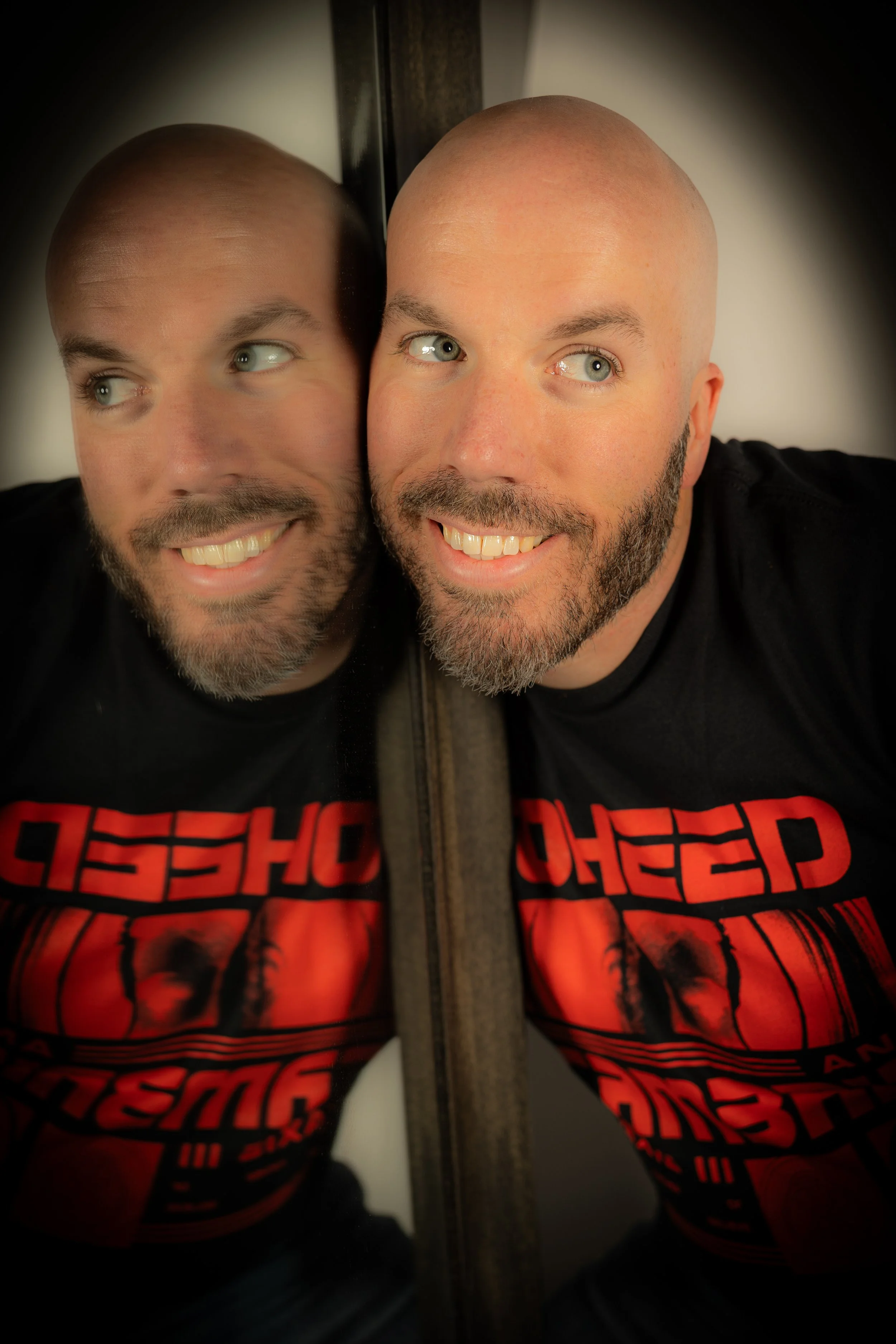 A man with a beard and a shaved head smiling while leaning against a mirror, with his reflection showing his face and part of his T-shirt that has red and black graphics.