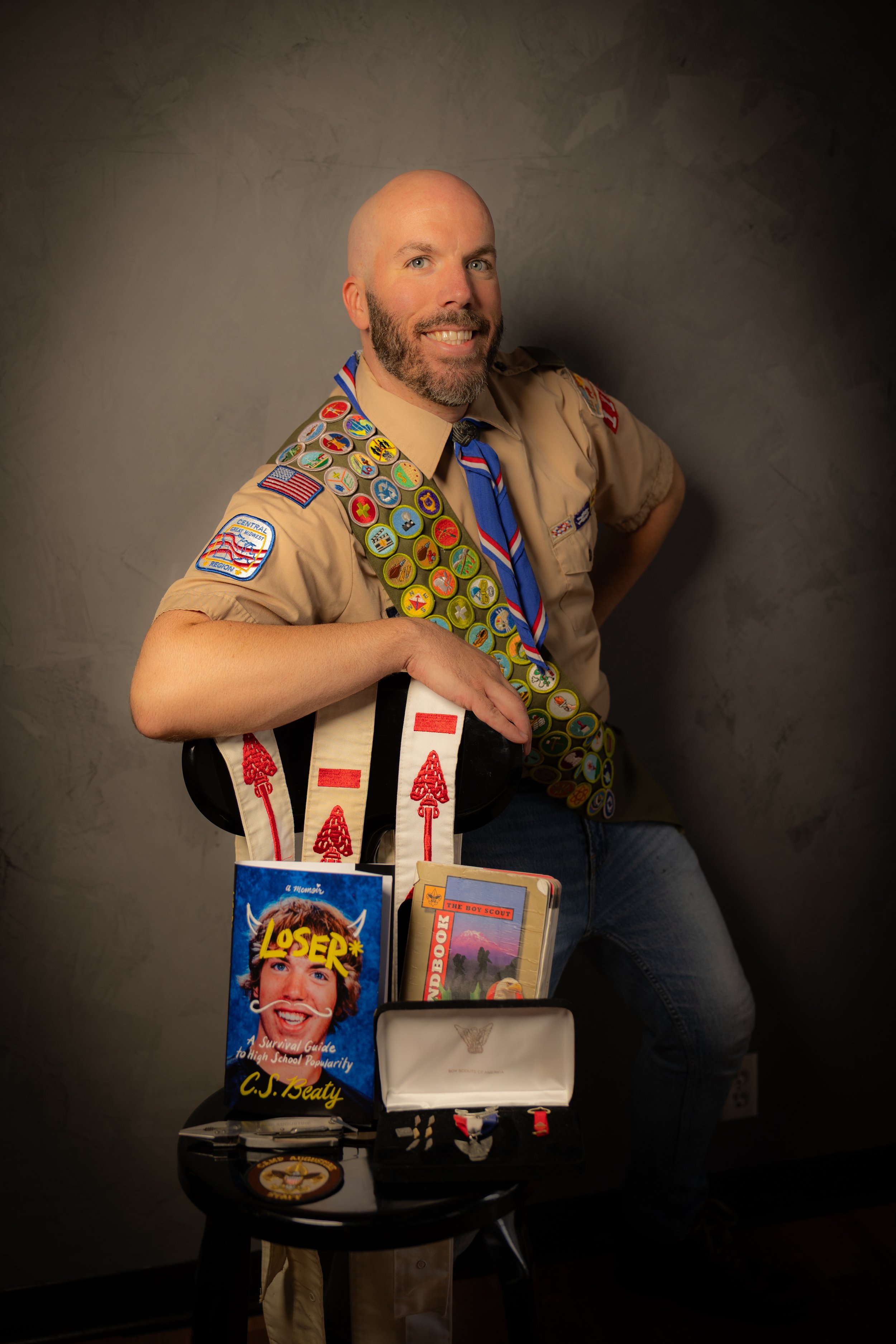 A man in a scout uniform with numerous patches and badges, wearing a neckerchief, smiling and posing next to a table with scout-related books, a medal, and scout awards.